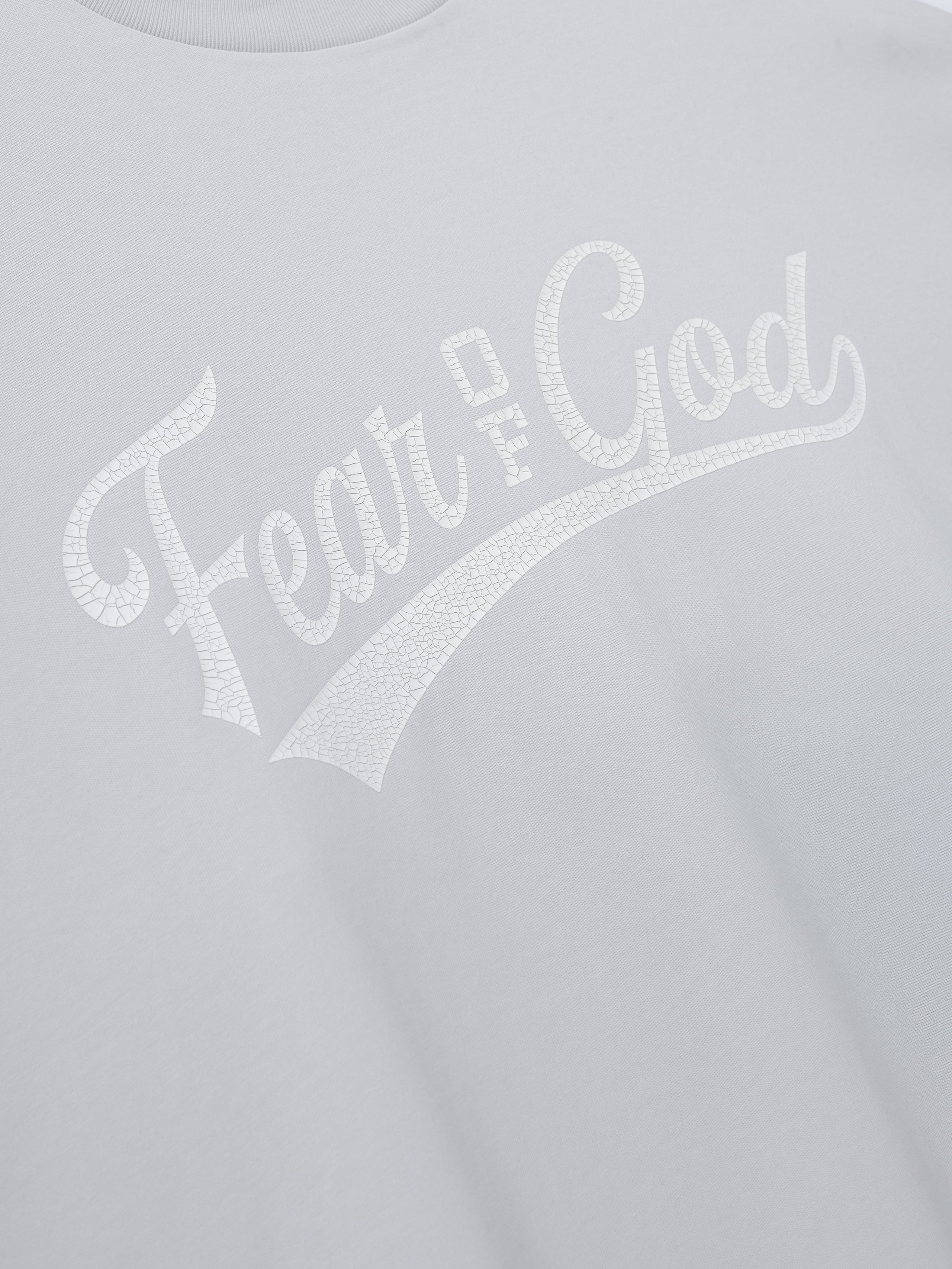 Merch Long Sleeve Tee in light grey featuring a cracked white "Fear of God" script printed across the chest on a soft casual top.