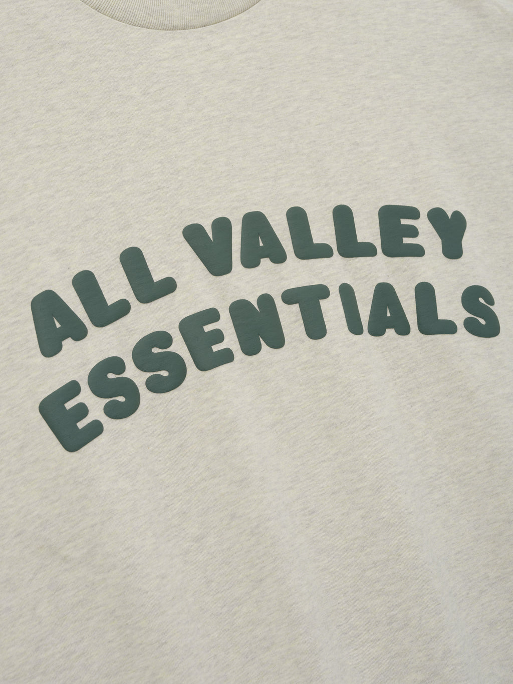 Merch Tee in light heathered neutral shade featuring soft fabric with bold dark green "ALL VALLEY ESSENTIALS" text curved across the front for a relaxed casual top.