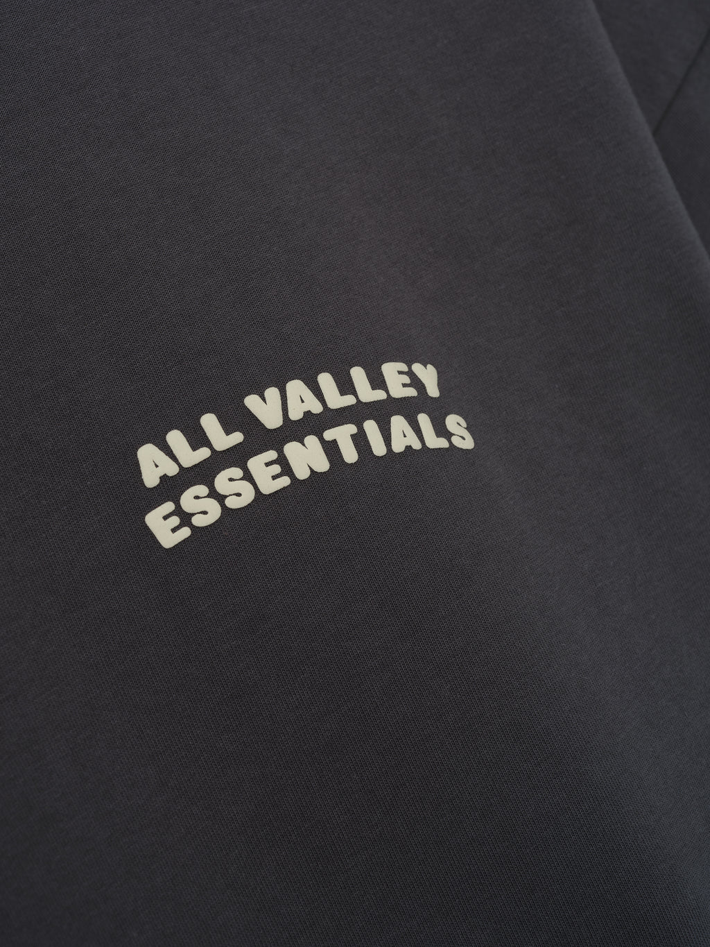 Merch Tee in deep iron grey featuring soft fabric with a minimalist curved cream "ALL VALLEY ESSENTIALS" text printed on the chest for a clean, casual top.