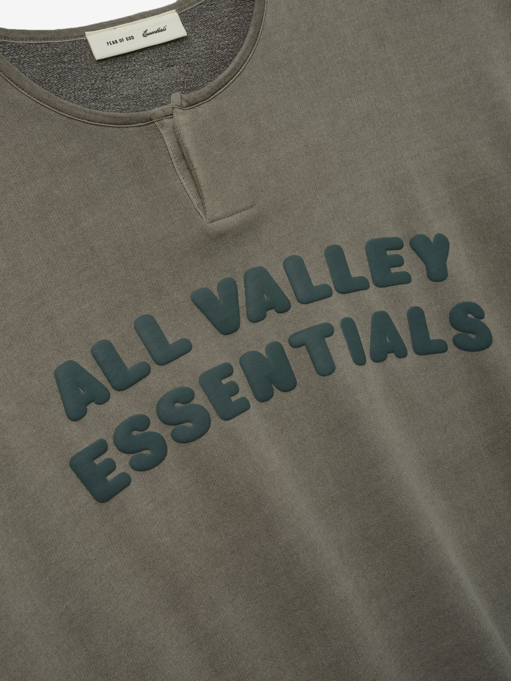 Close-up of the front upper section of a 3/4 Henley Sweatshirt in washed seal color, featuring a small buttoned placket and raised green lettering reading "ALL VALLEY ESSENTIALS".