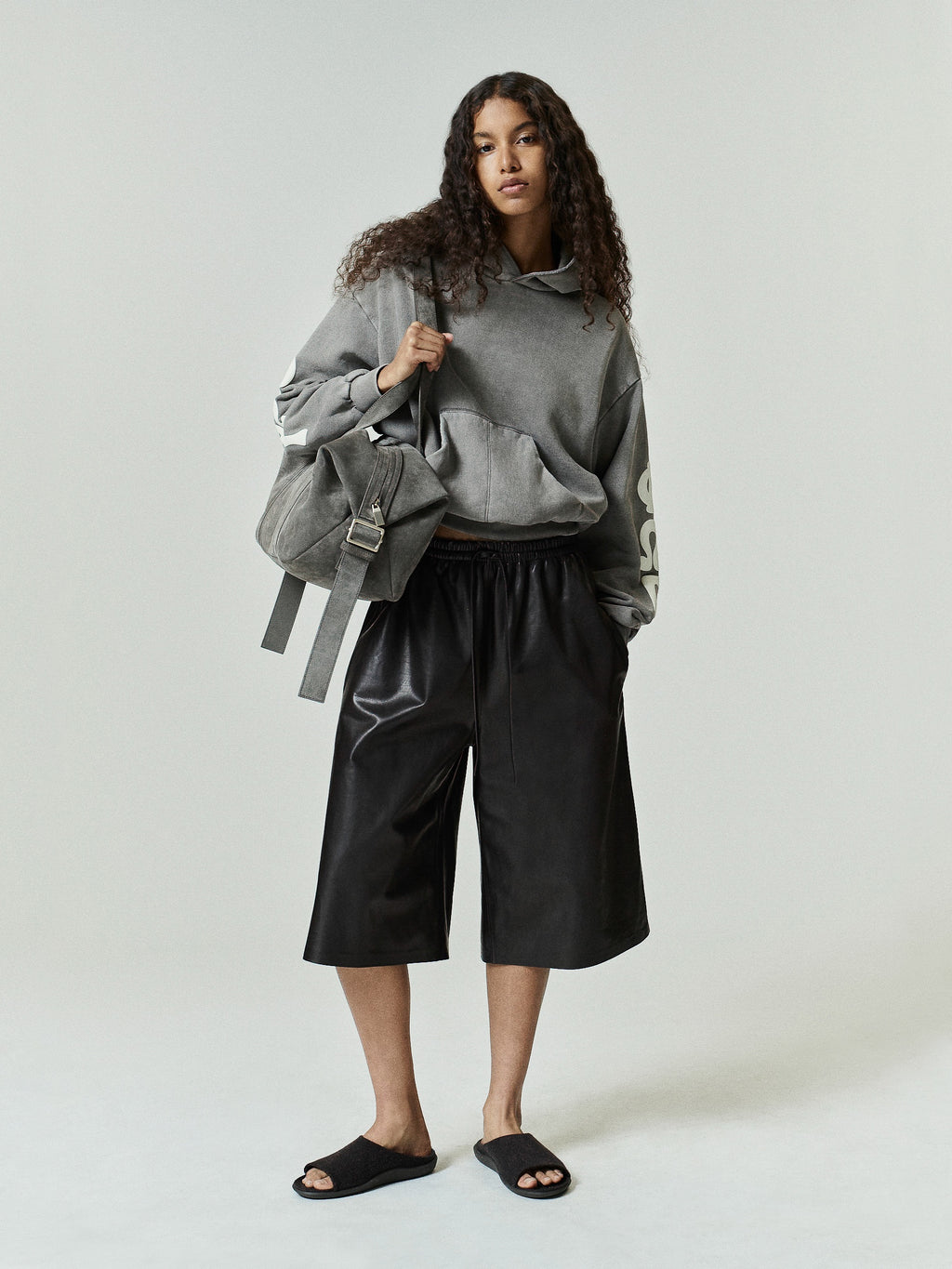 Womens 90s Fleece Hoodie in washed seal green with a relaxed fit, featuring a front kangaroo pocket and bold cream graphic lettering on the sleeve, styled with black wide-leg shorts.