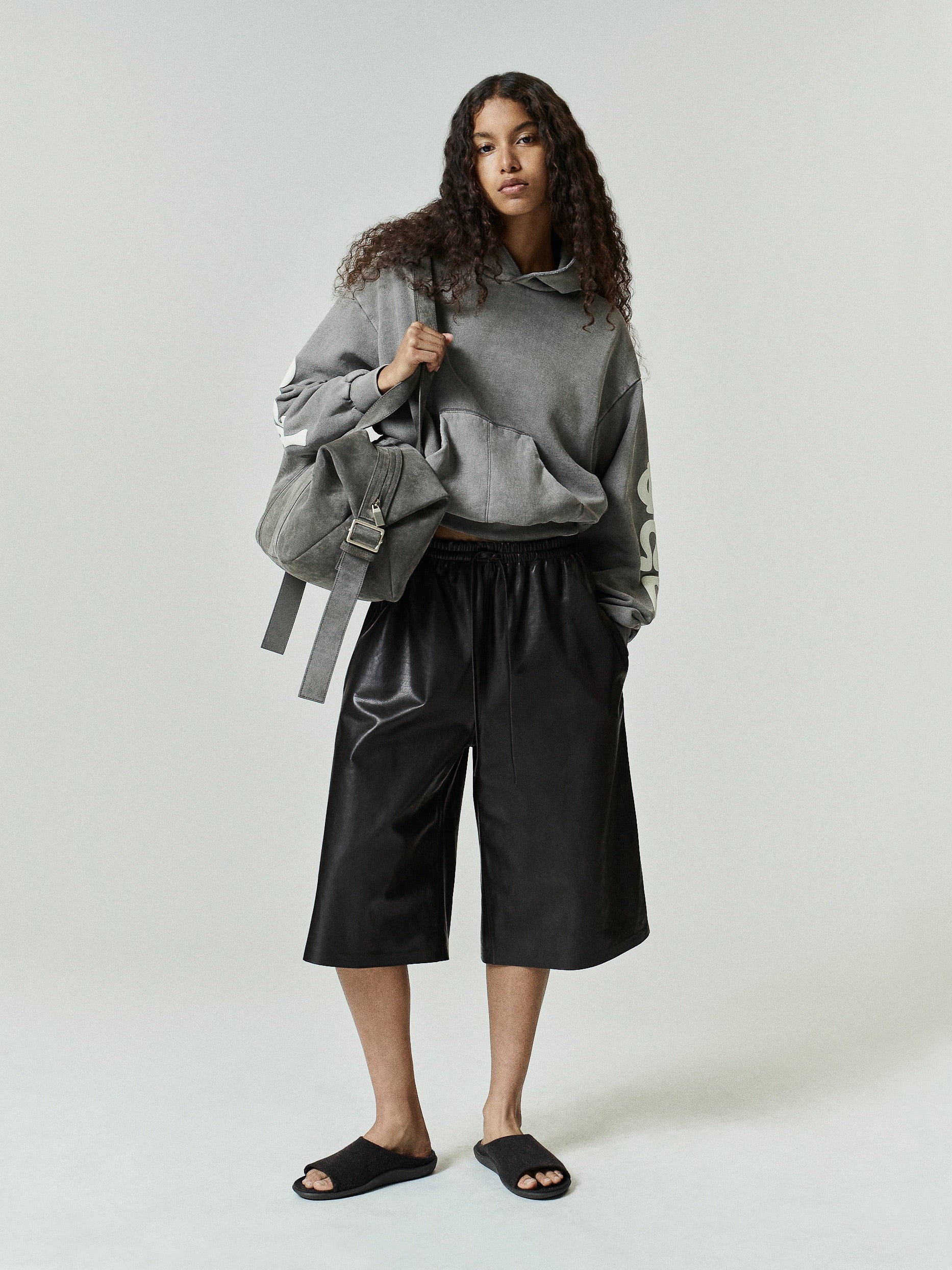 Womens 90s Fleece Hoodie in washed seal green with a relaxed fit, featuring a front kangaroo pocket and bold cream graphic lettering on the sleeve, styled with black wide-leg shorts.