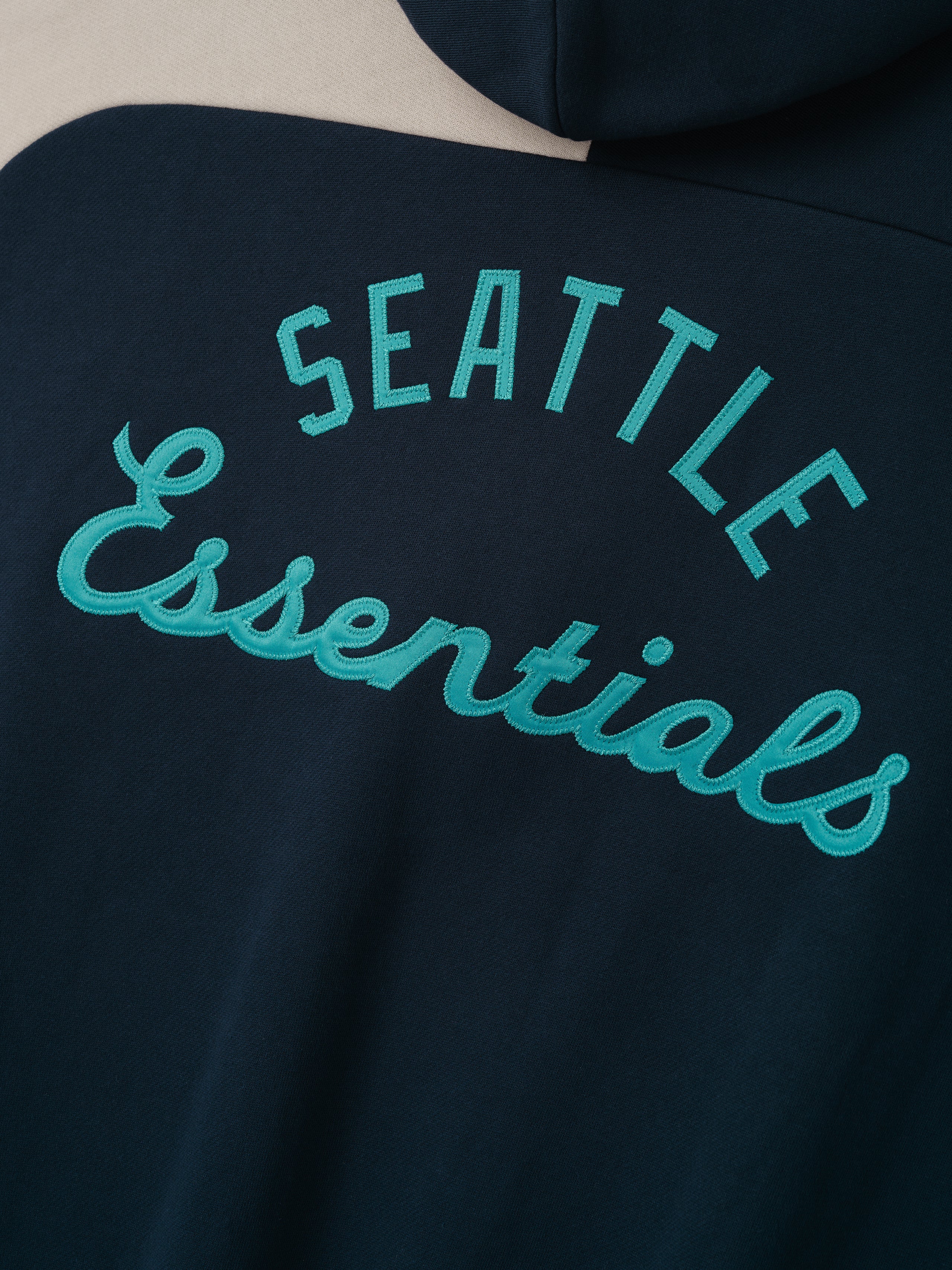 Close-up of the back of the Mariners Raglan Half Zip Sport Hoodie showing teal embroidered "Seattle Essentials" lettering on dark sapphire fabric with a cream-colored accent.