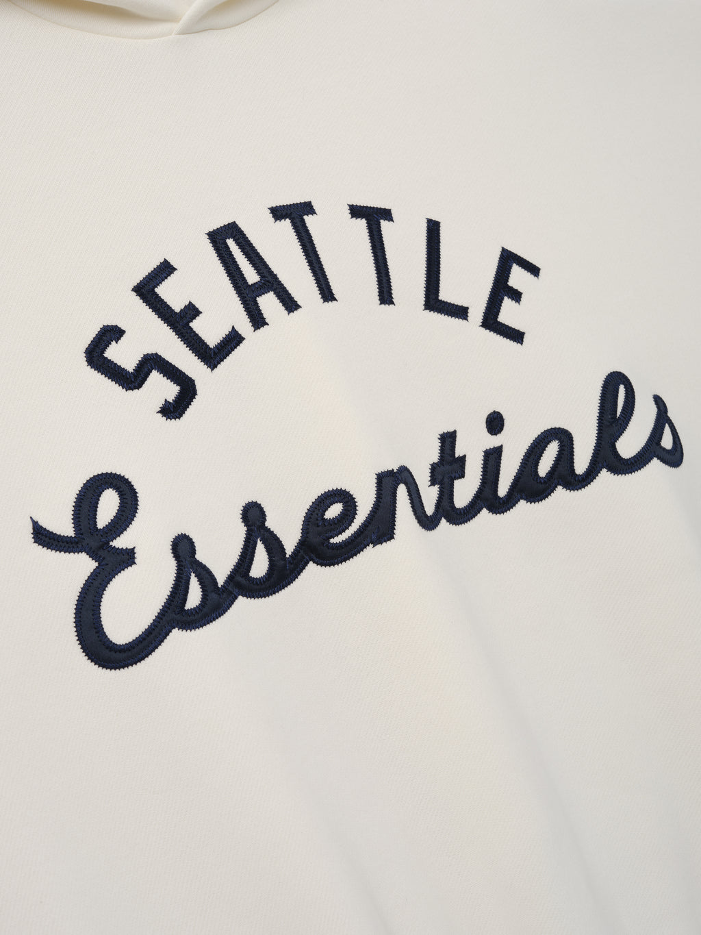 Close-up of the front of the Mariners Sport Hoodie in soft crema white fabric, featuring raised navy embroidered "Seattle Essentials" text in a bold, classic font.