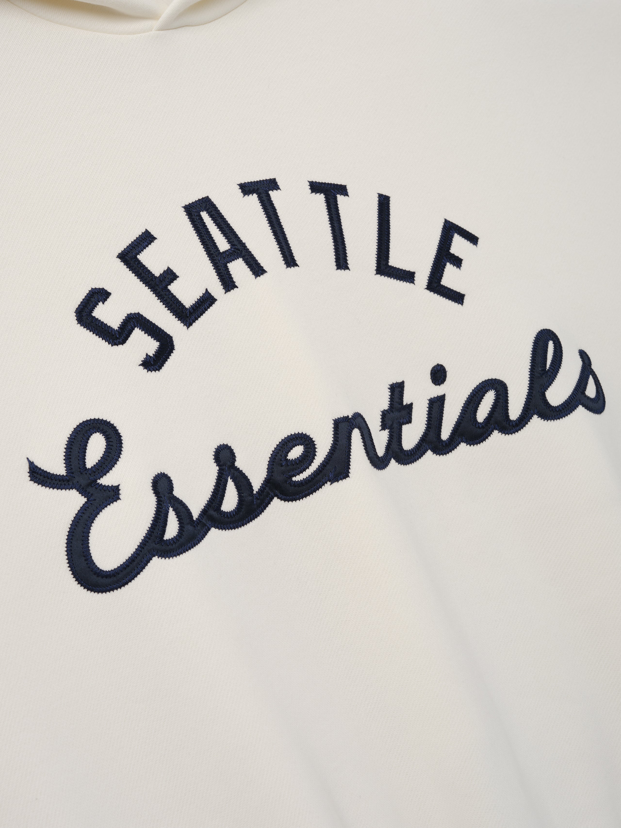 Close-up of the front of the Mariners Sport Hoodie in soft crema white fabric, featuring raised navy embroidered "Seattle Essentials" text in a bold, classic font.