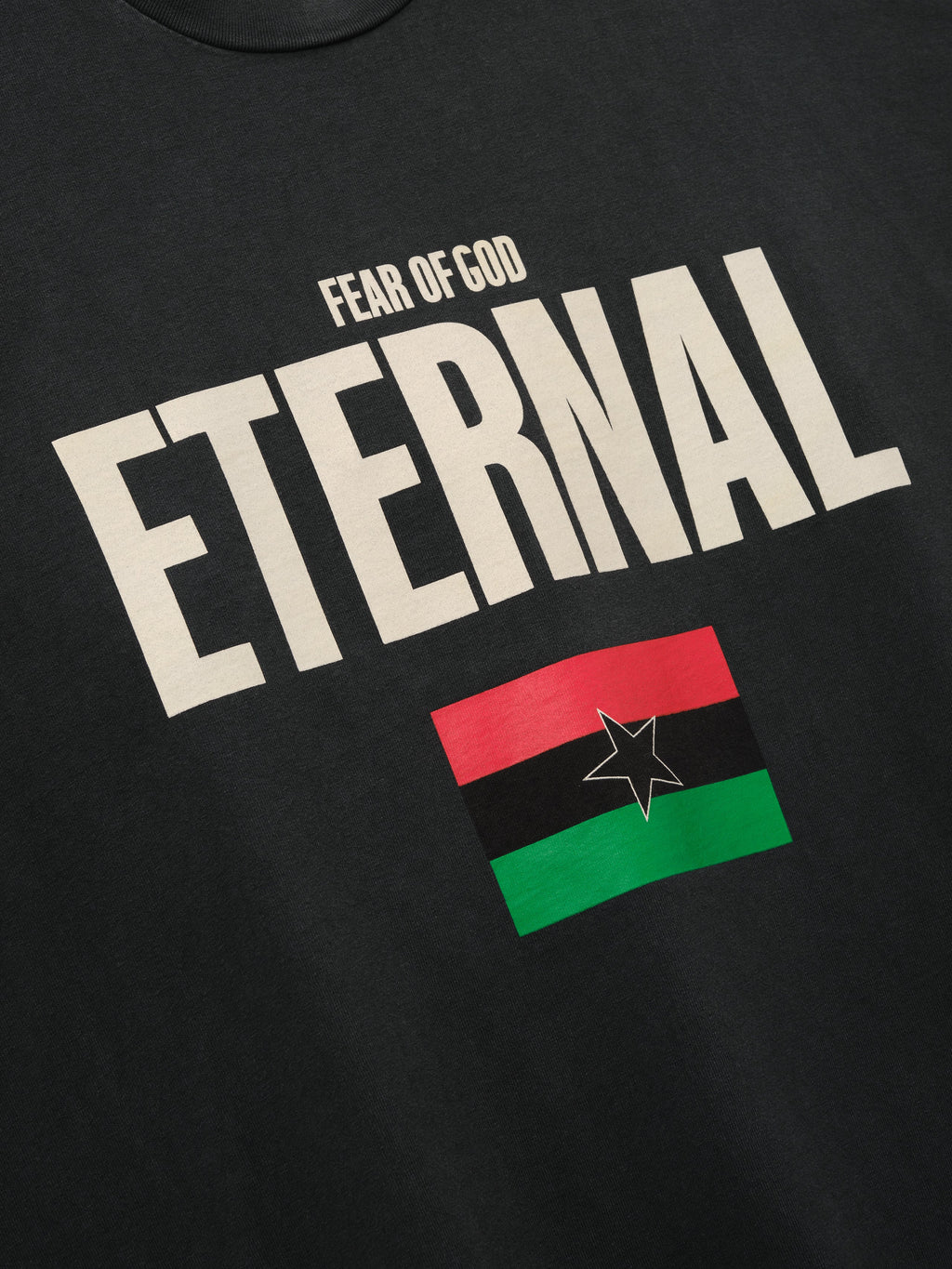 Reflection Eternal Tee in black shown in close detail with cream Fear of God Eternal lettering and red green black flag with star as Tops