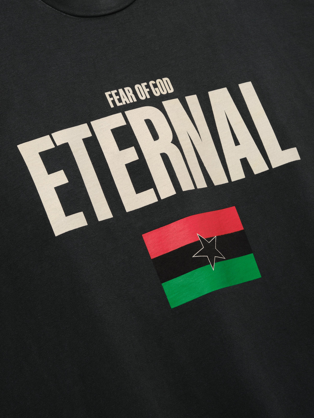 Reflection Eternal Tee in black shown in close detail with cream Fear of God Eternal lettering and red green black flag with star as Tops