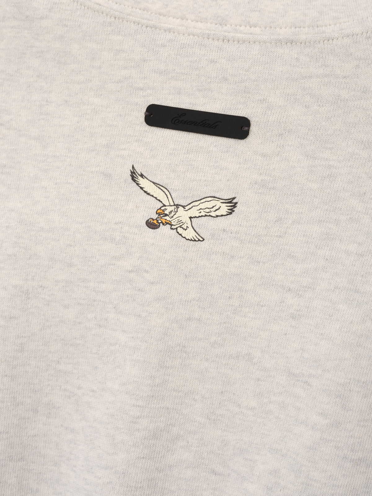Eagles Merch Tee cream fabric close up with embroidered flying eagle emblem and black chest label in a detailed Tops product view