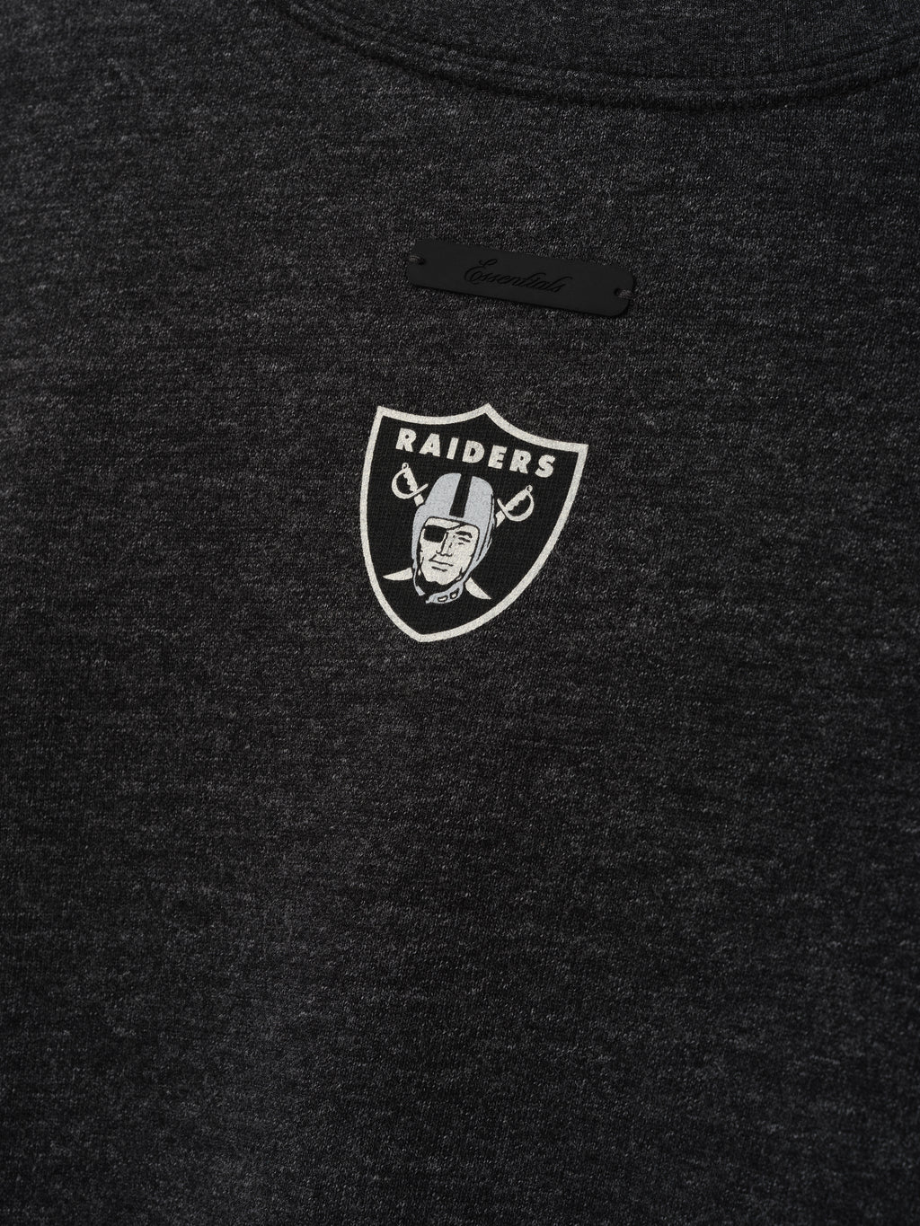 Raiders Merch Tee charcoal fabric close up featuring a small white Raiders shield emblem and black chest label in a clean Tops product detail view