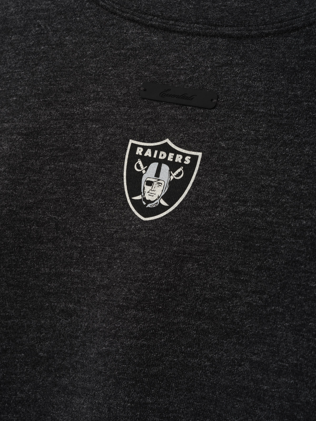 Raiders Merch Tee charcoal fabric close up featuring a small white Raiders shield emblem and black chest label in a clean Tops product detail view