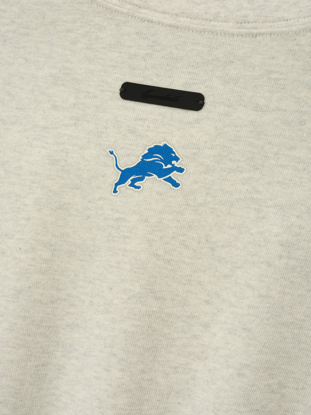 Lions Merch Tee cream fabric close up featuring an embroidered blue lion emblem and black chest label in a detailed Tops product view