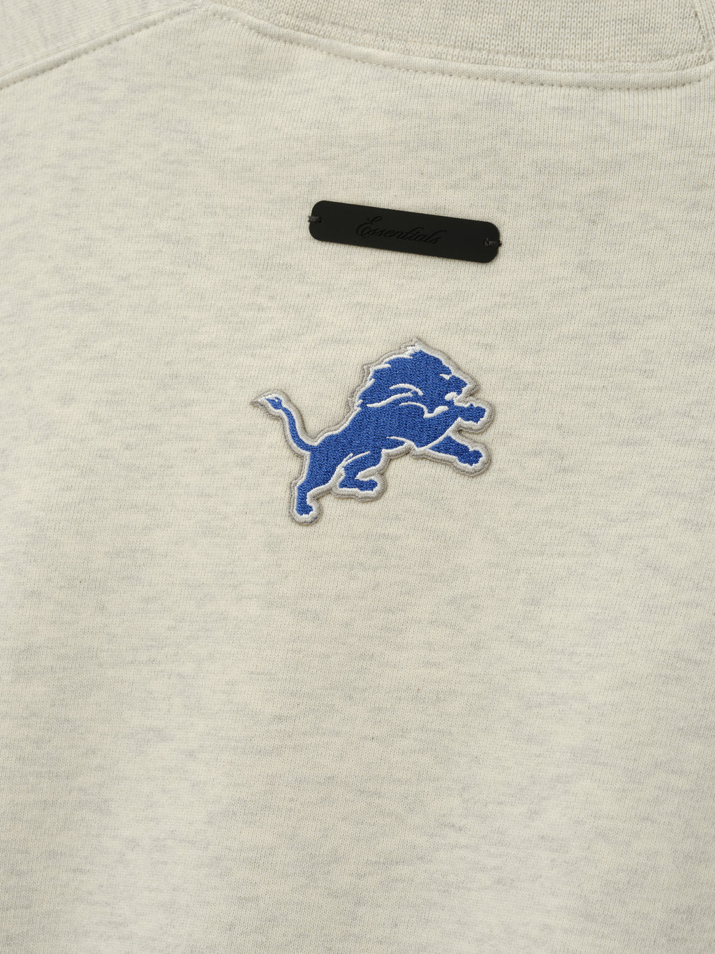 Lions 90s Relaxed Crewneck cream sweatshirt close up showing embroidered blue Detroit Lions logo and black chest label as a Tops product detail