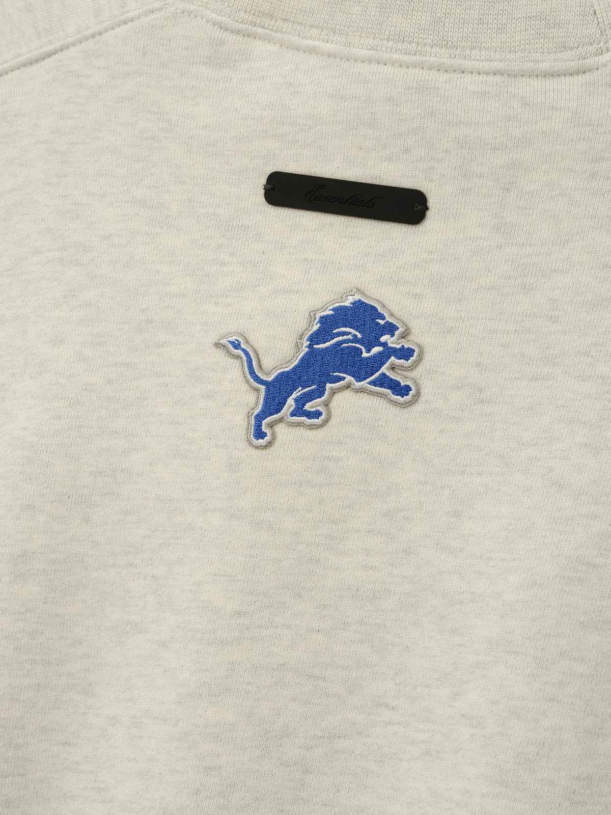 Lions 90s Relaxed Crewneck cream sweatshirt close up showing embroidered blue Detroit Lions logo and black chest label as a Tops product detail