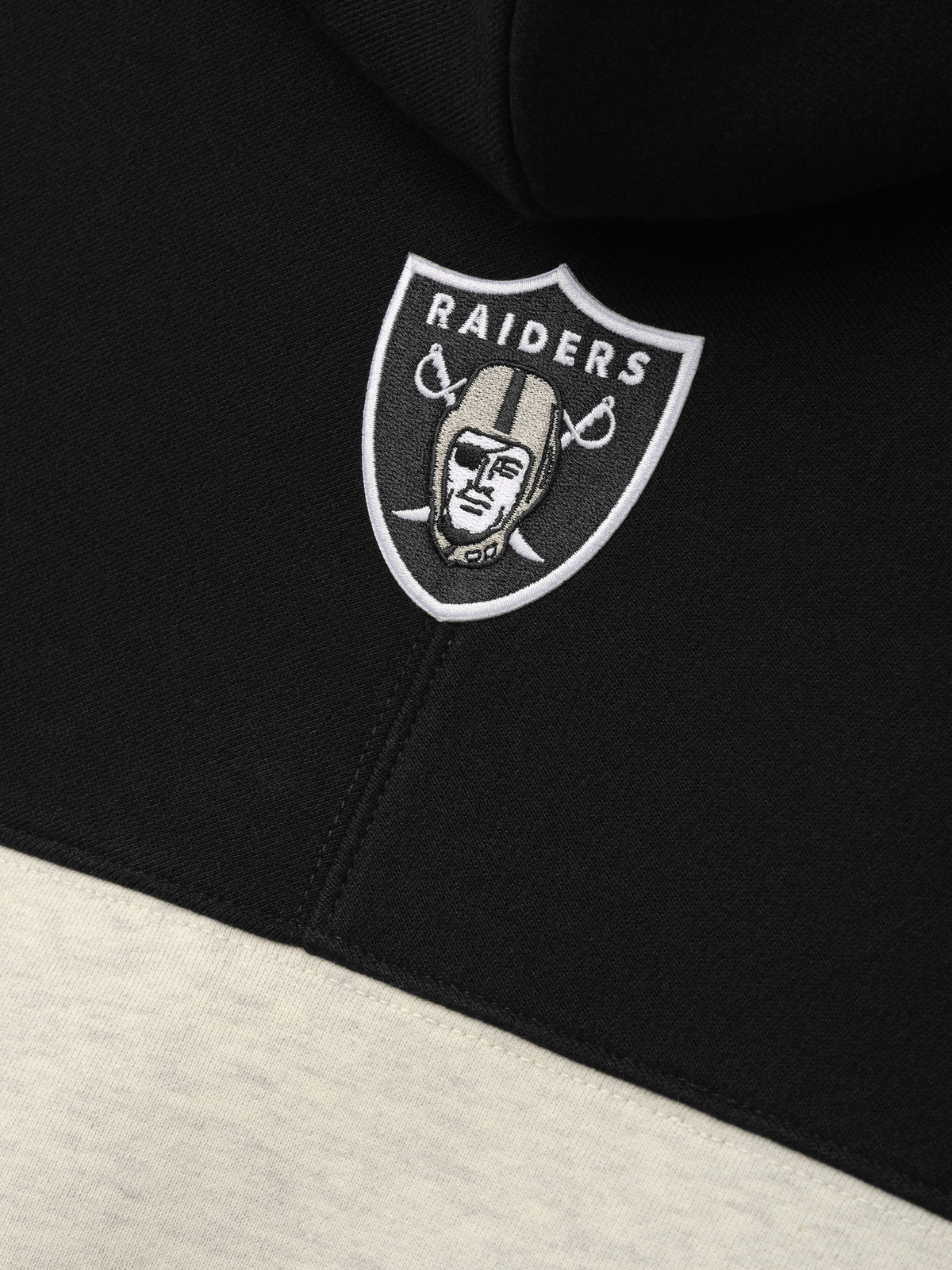 Raiders 90s Relaxed Hoodie close up showing an embroidered Raiders shield logo on black fabric above a cream color block as a Tops product detail