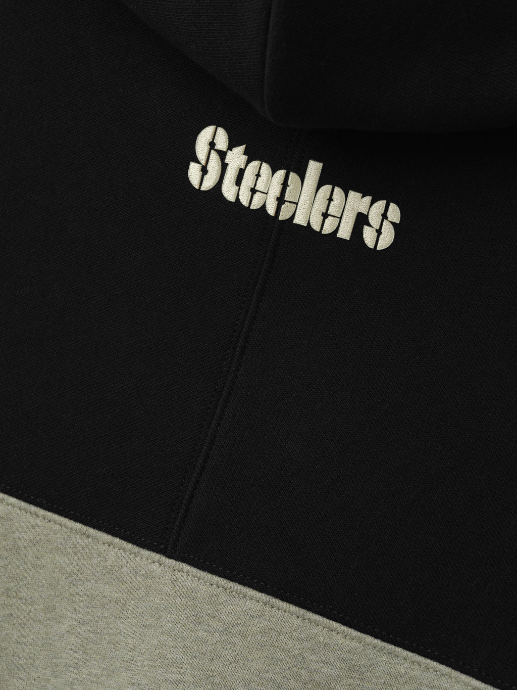Steelers 90s Relaxed Hoodie close up showing cream Steelers lettering on black fleece above a gray color blocked panel as a Tops product detail