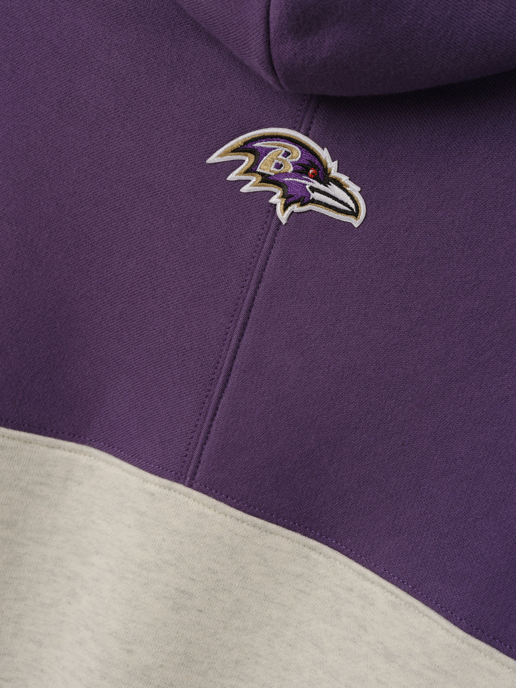 Ravens 90s Relaxed Hoodie close up with embroidered Ravens logo on purple fleece above a cream color blocked panel as a Tops product detail