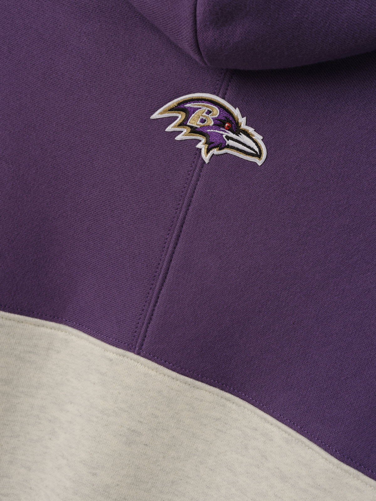 Ravens 90s Relaxed Hoodie close up with embroidered Ravens logo on purple fleece above a cream color blocked panel as a Tops product detail