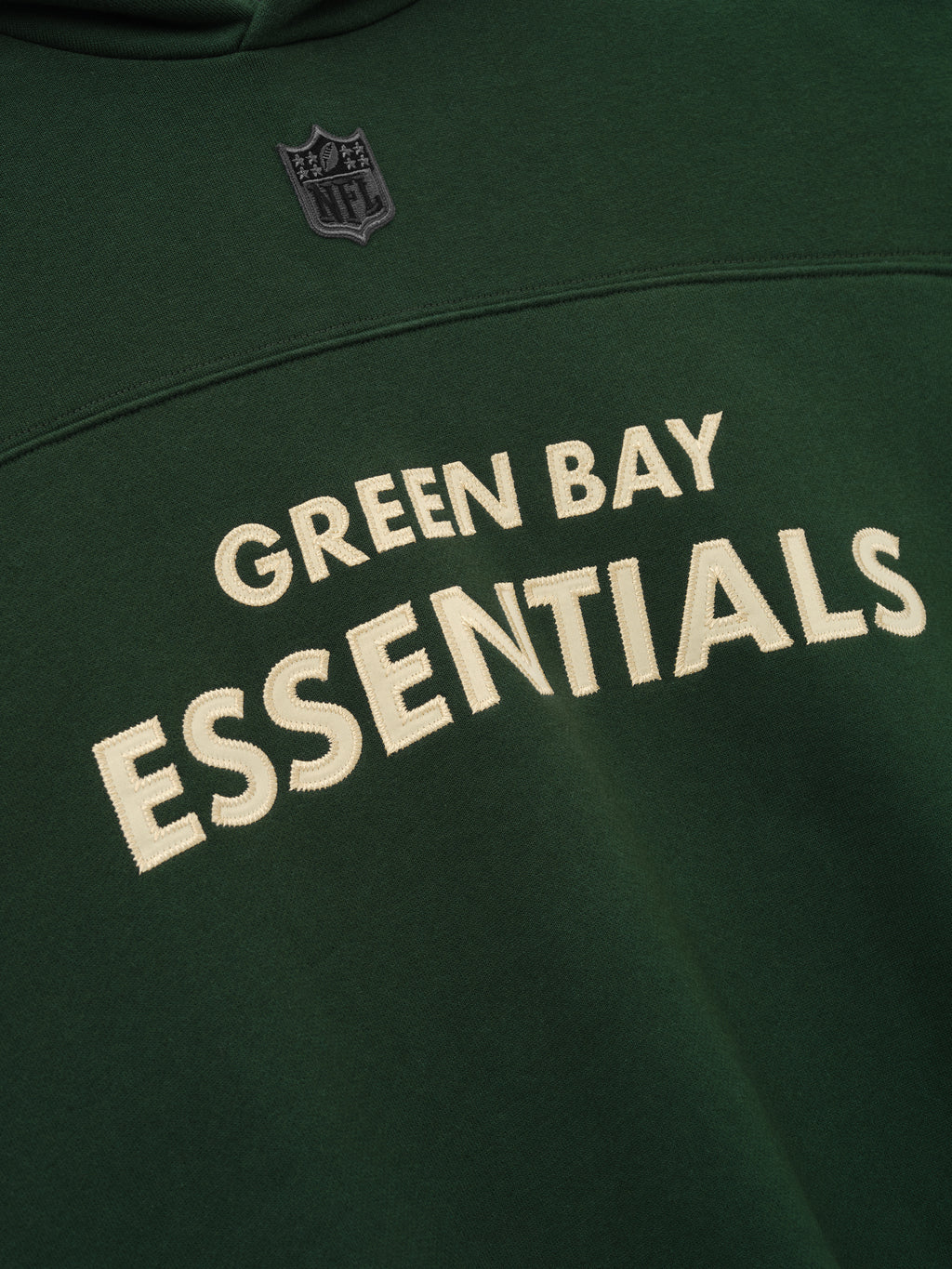 Packers 90s Relaxed Hoodie close up showing Green Bay Essentials lettering and NFL shield embroidered on dark green fleece as a Tops product detail