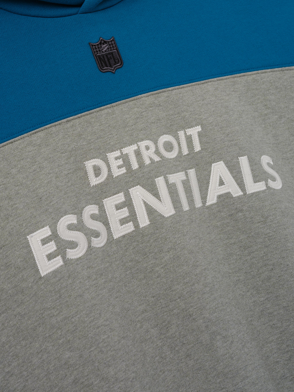 Lions 90s Relaxed Hoodie close up featuring Detroit Essentials lettering and NFL shield on blue and gray color blocked fleece as a Tops product detail
