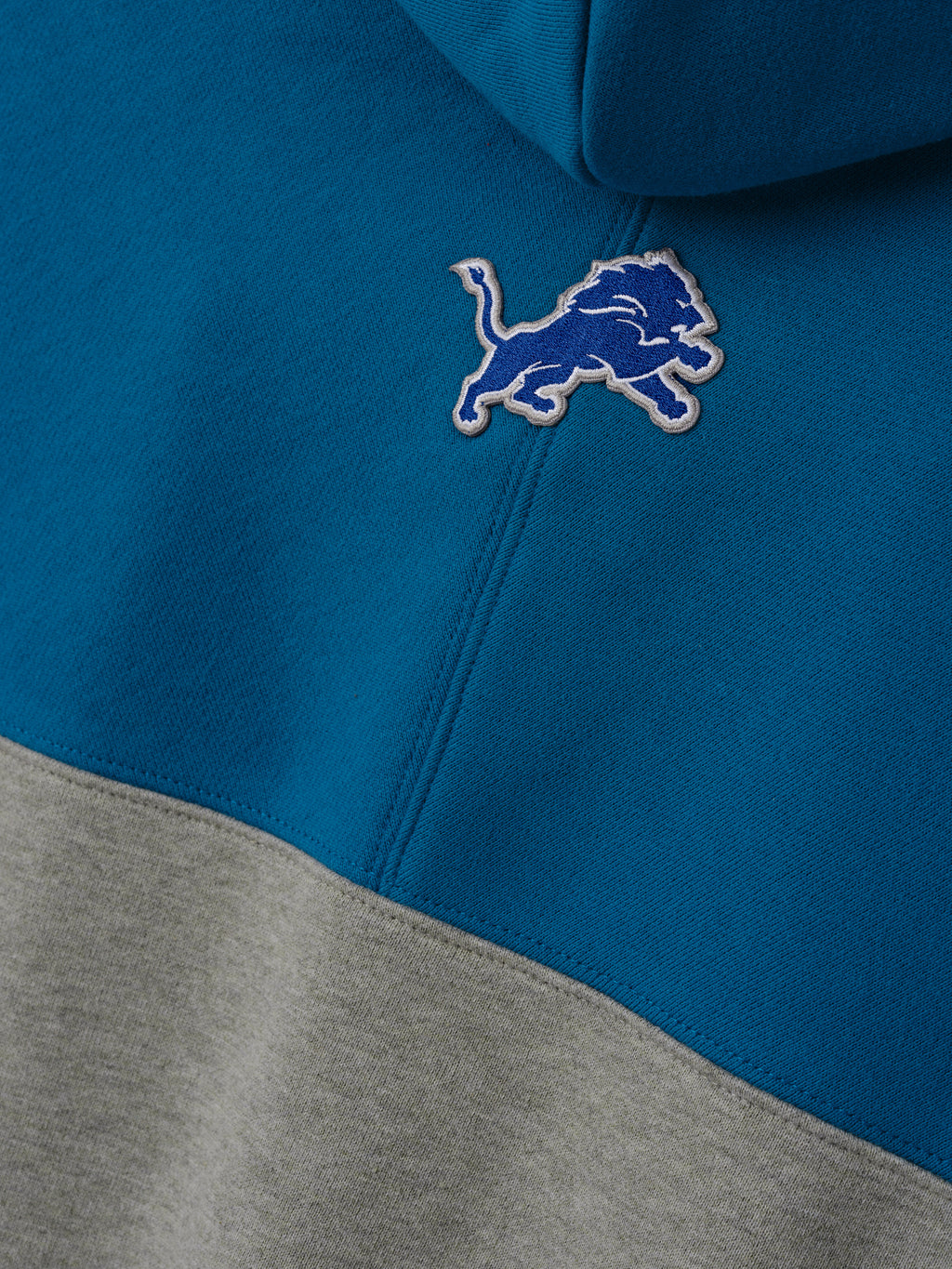 Lions 90s Relaxed Hoodie close up showing an embroidered Detroit Lions logo on blue and gray color blocked fleece with a stitched seam as a Tops product detail