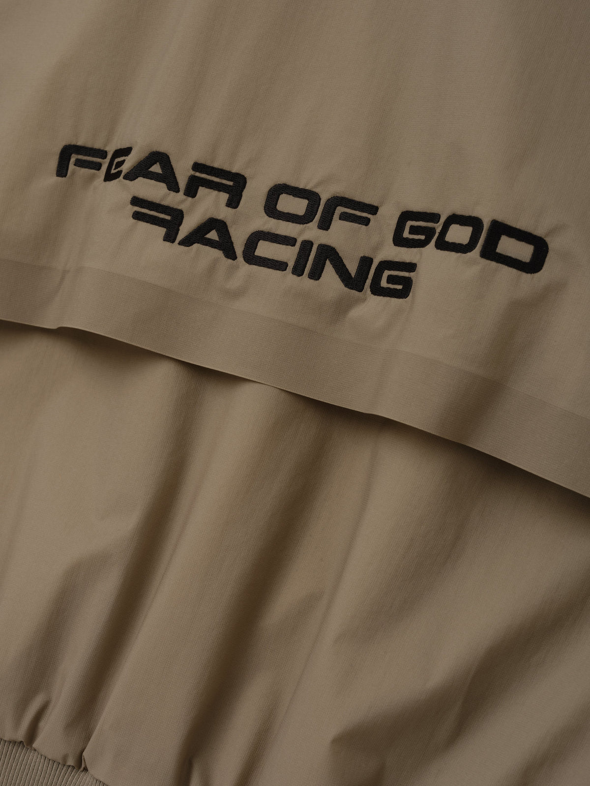 Womens Pullover Track Jacket in taupe with black Fear of God Racing branding on a layered panel and elastic hem shown in close detail as Outerwear