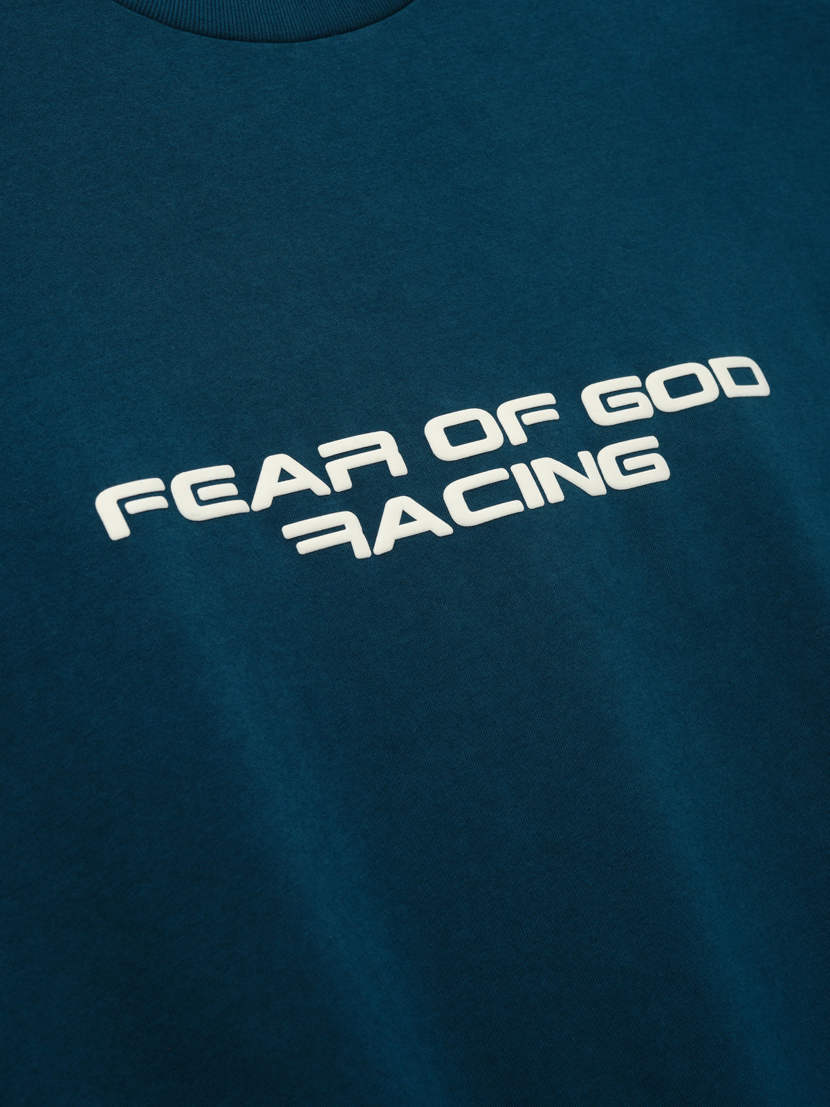Merch Tee in dark teal with cream Fear of God Racing chest graphic and crew neckline shown in close up as Tops
