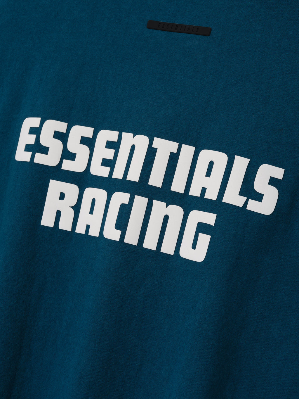Kids Merch Tee in deep teal featuring a bold white Essentials Racing graphic across the back with a small neck label shown in close detail as Tops