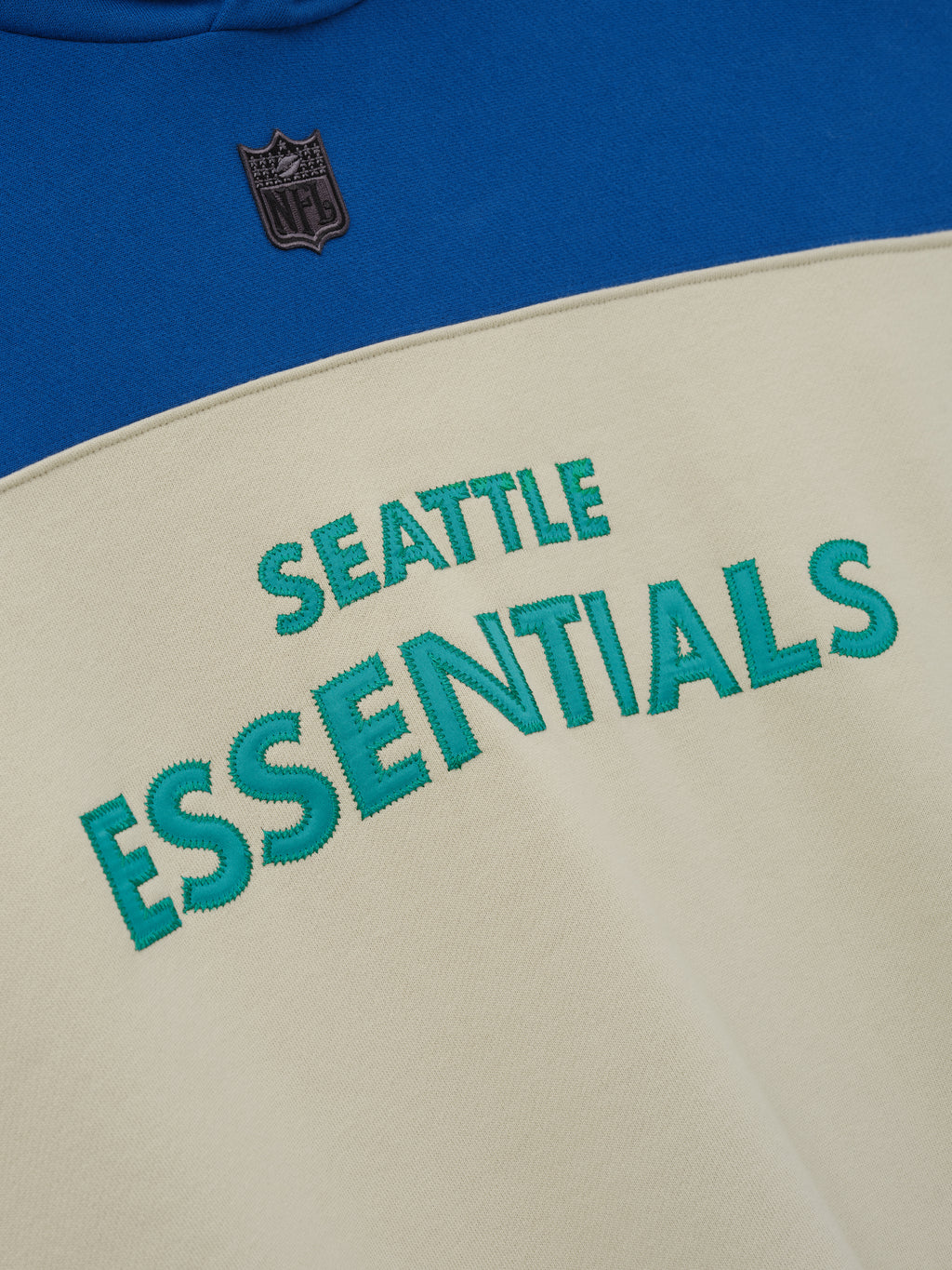 Seahawks 90s Relaxed Hoodie close up in blue and cream fleece with Seattle Essentials lettering and NFL shield detail shown as a Tops product view