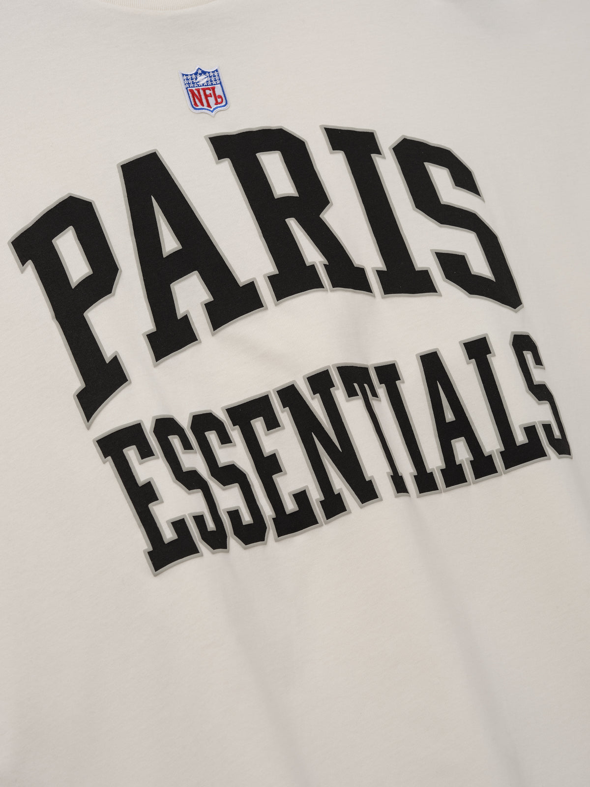 NFL Paris Merch Tee in off white featuring an NFL shield and bold black PARIS ESSENTIALS graphic lettering across the front close up Tops