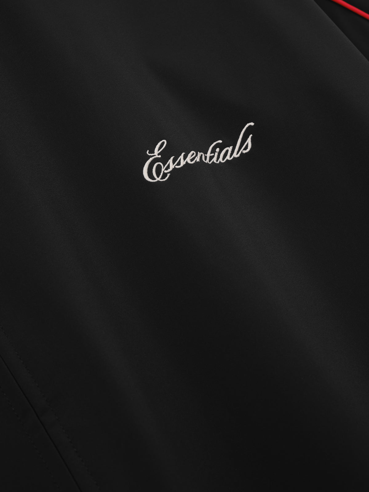 Marathon Modern Track Jacket in black with white Essentials script logo and red piping detail shown in close up as Outerwear