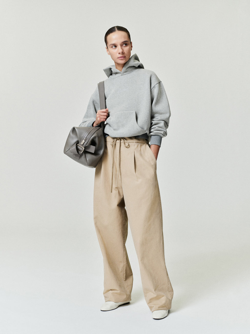 Person wearing a gray hoodie and beige pants holding a gray bag on a white background