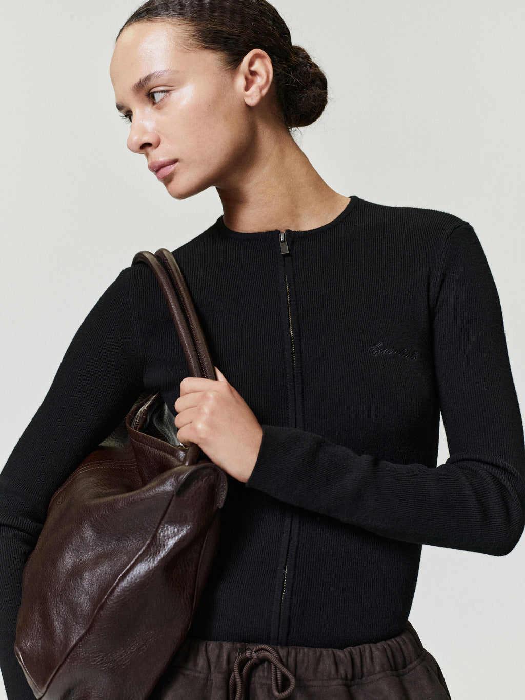 Woman holding a brown leather bag wearing a black jacket with a brand logo.