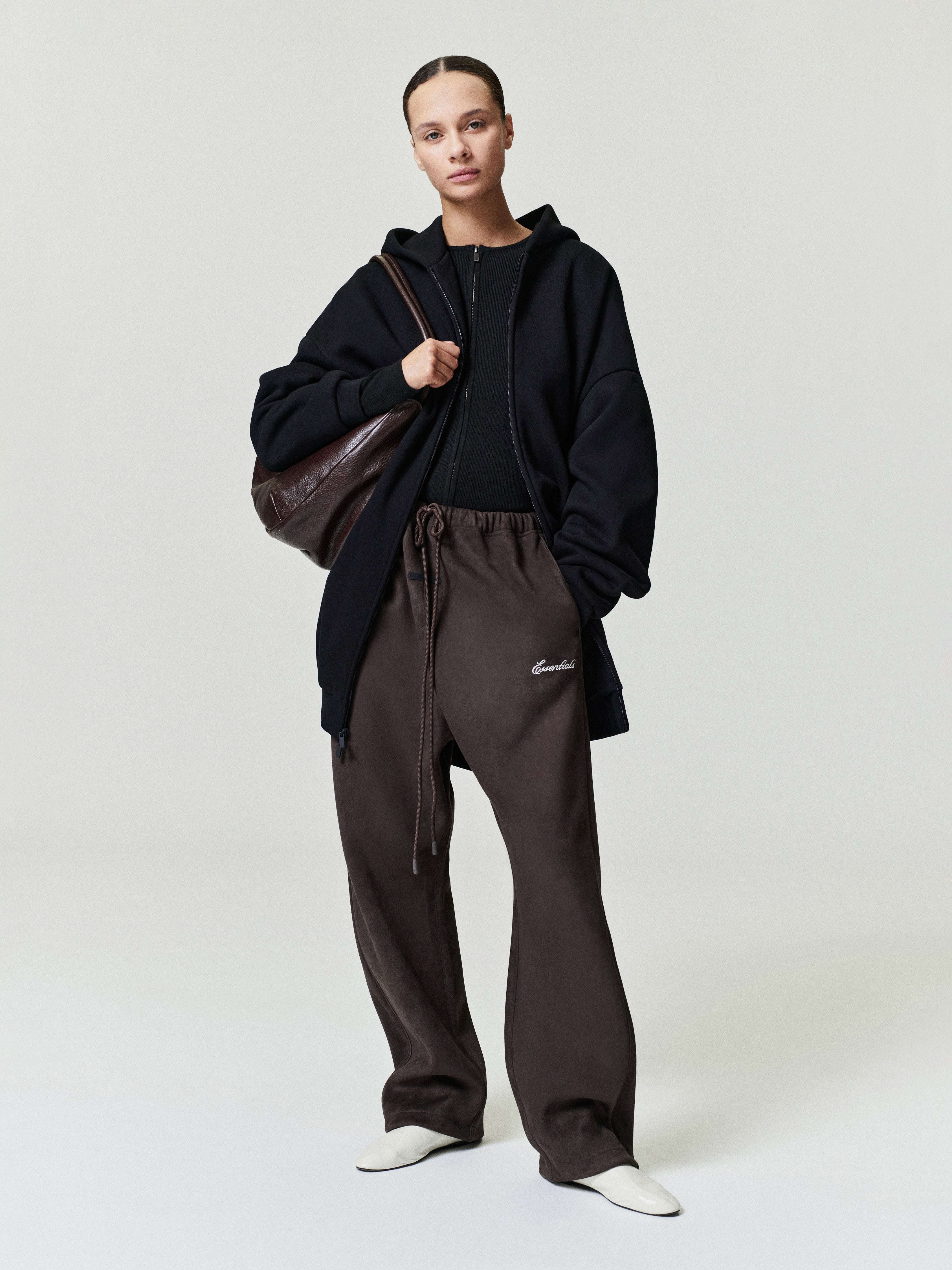Person wearing a black hoodie and brown pants with a brown bag on a white background
