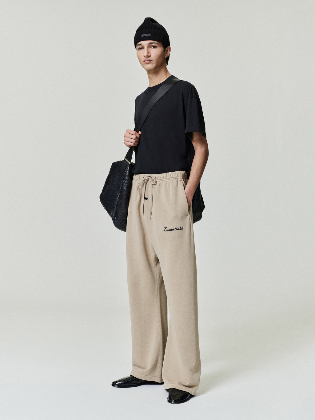 Person wearing a black top and beige essentials flare sweatpants with essentials script puff print logo , holding a black bag on a light gray background