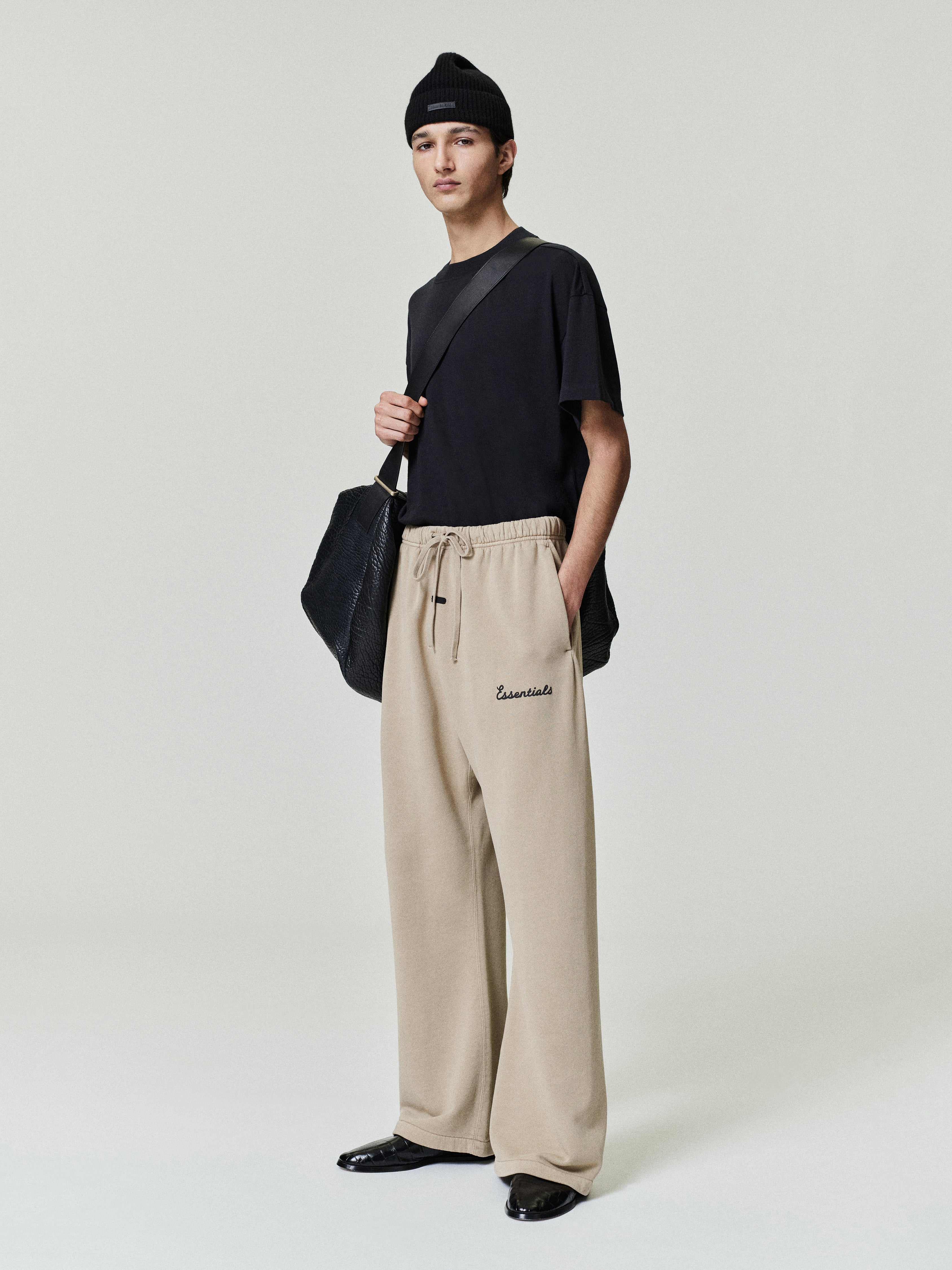 Person wearing a black top and beige essentials flare sweatpants with essentials script puff print logo , holding a black bag on a light gray background