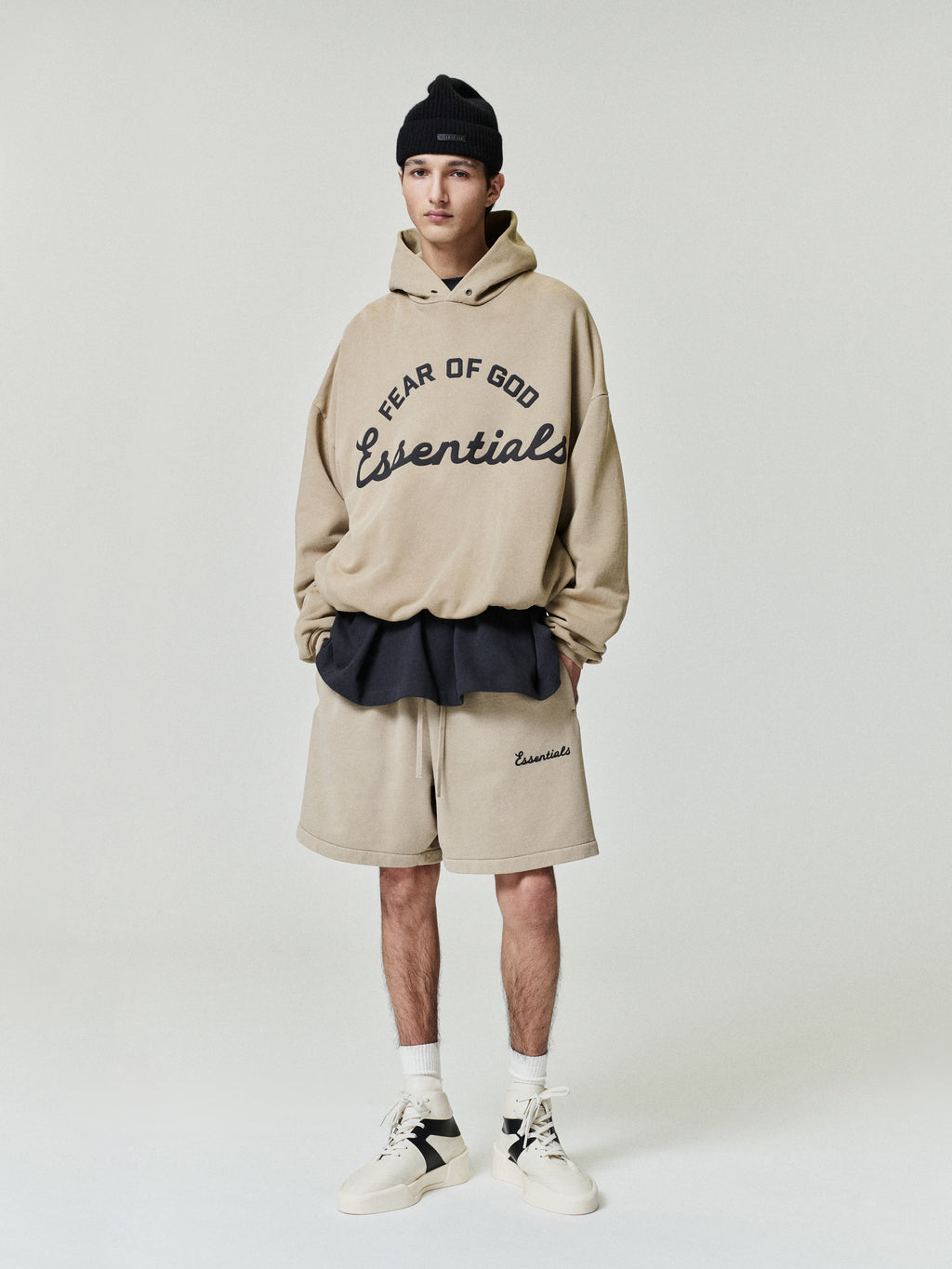 Person wearing a beige 'Fear of God Essentials' hoodie and shorts on a white background