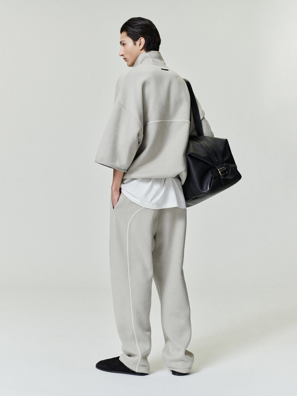 man wearing light beige cropped volume polo sweatshirt with 3/4 sleeves and matching relaxed fleece pants carrying black leather bag