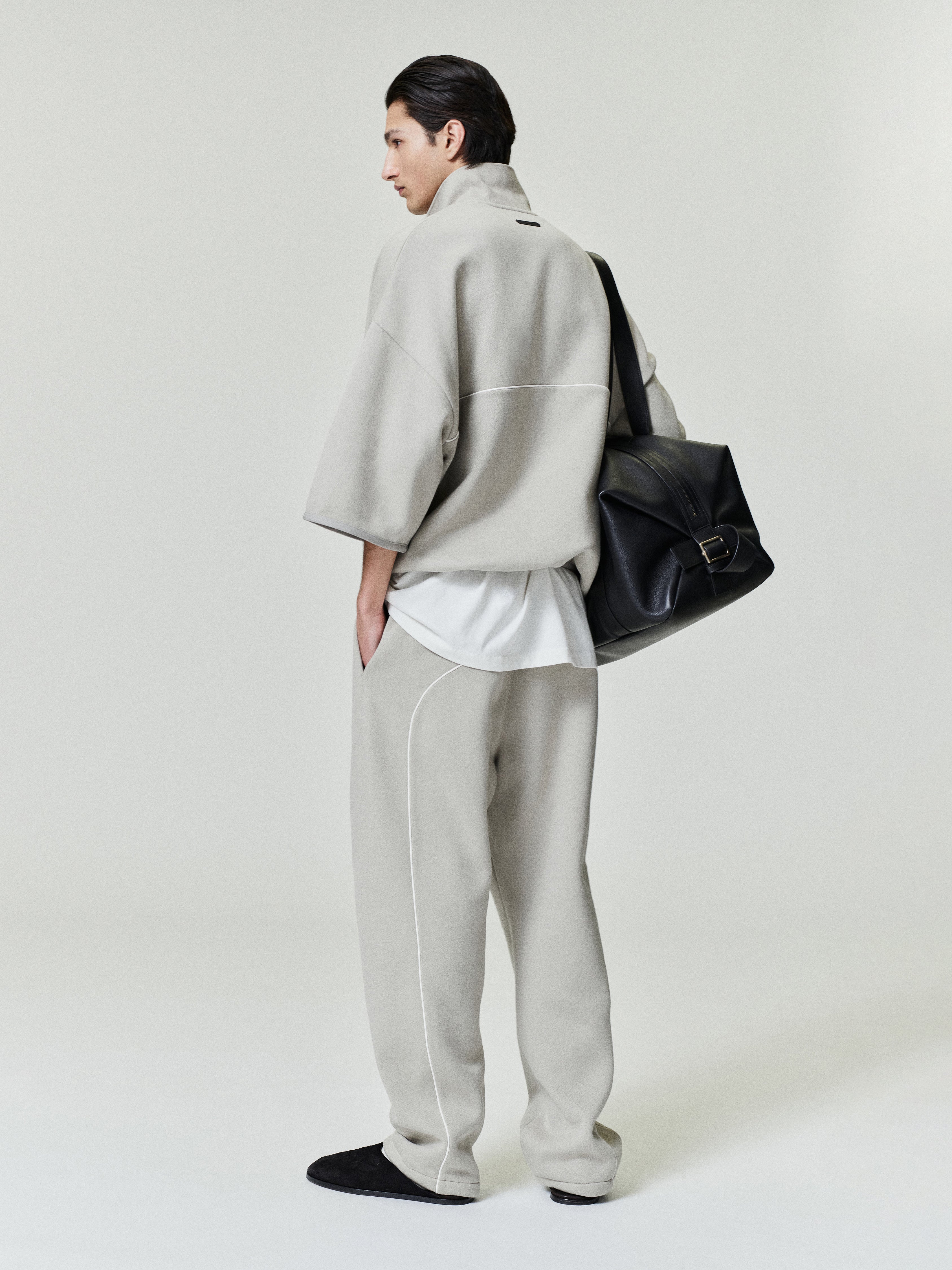 man wearing light beige cropped volume polo sweatshirt with 3/4 sleeves and matching relaxed fleece pants carrying black leather bag