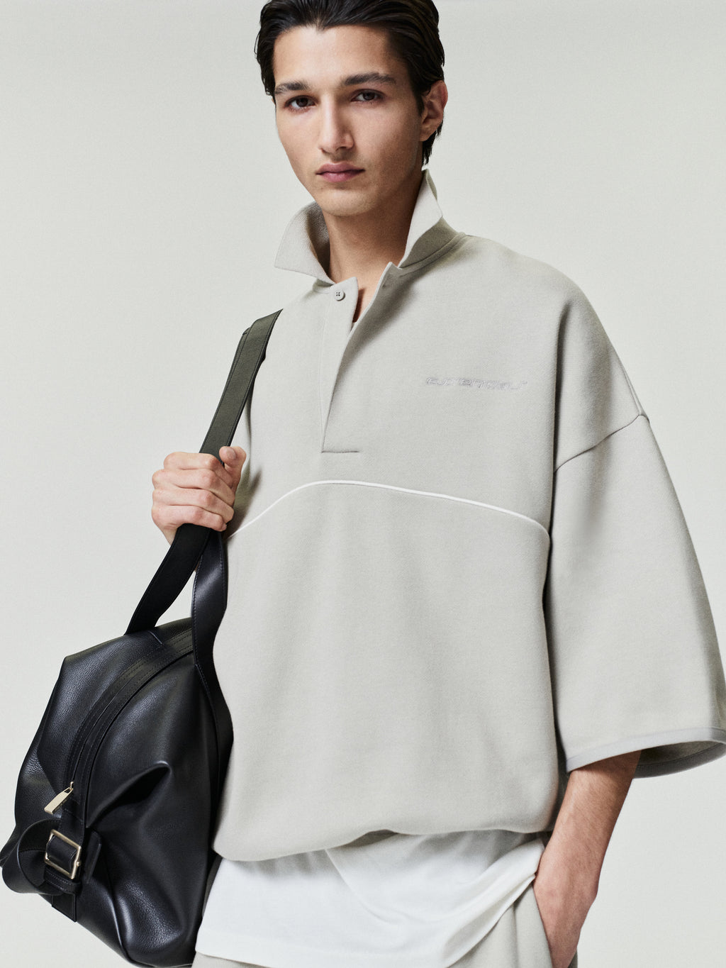 man wearing light beige cropped polo sweatshirt with 3/4 sleeves and subtle embroidered logo holding black leather bag
