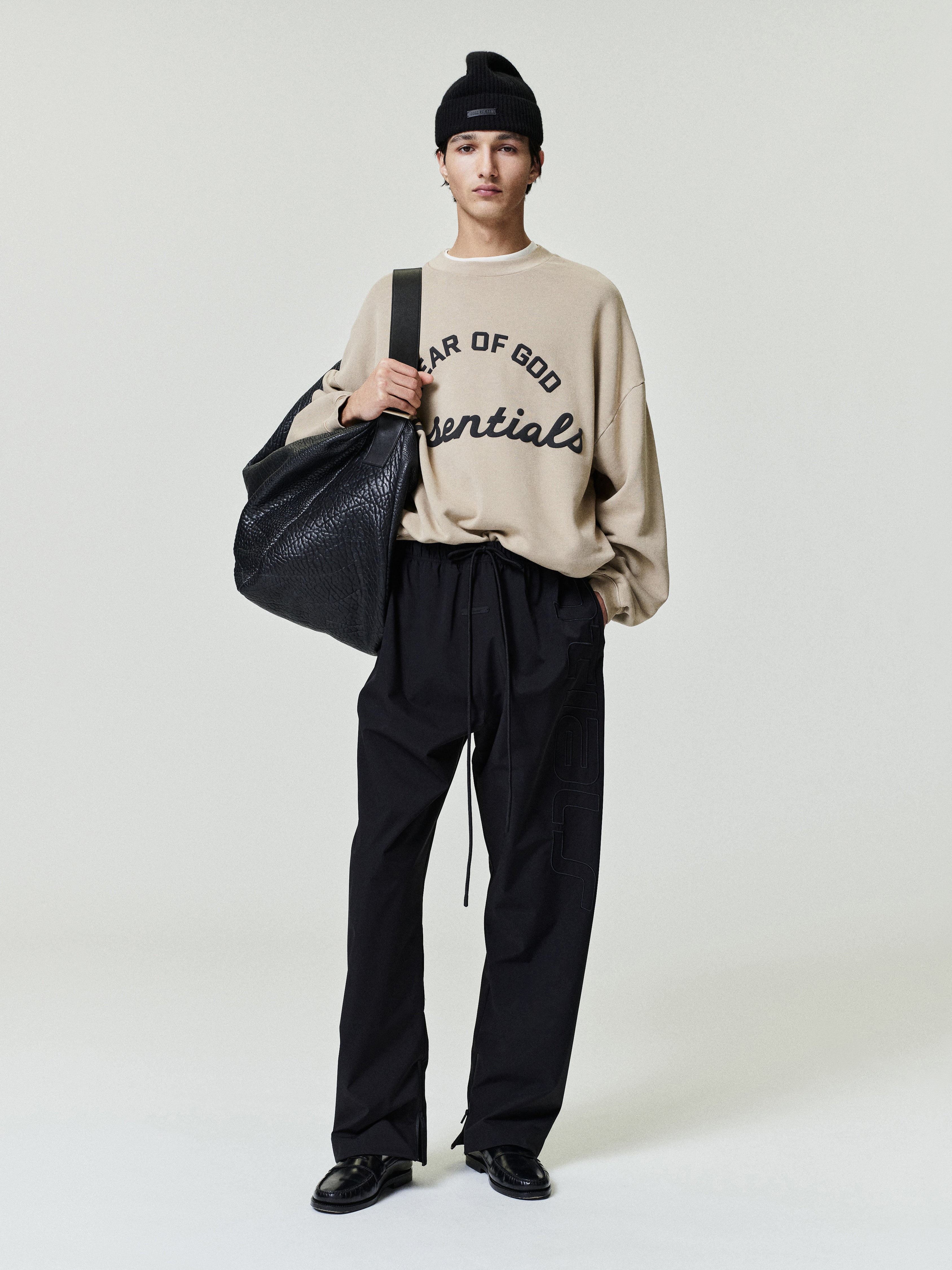 Person wearing a beige sweatshirt with text, black pants, and holding a black bag on a white background