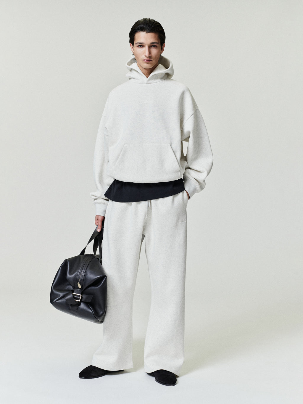 Person wearing a white hoodie and pants holding a black bag on a light gray background