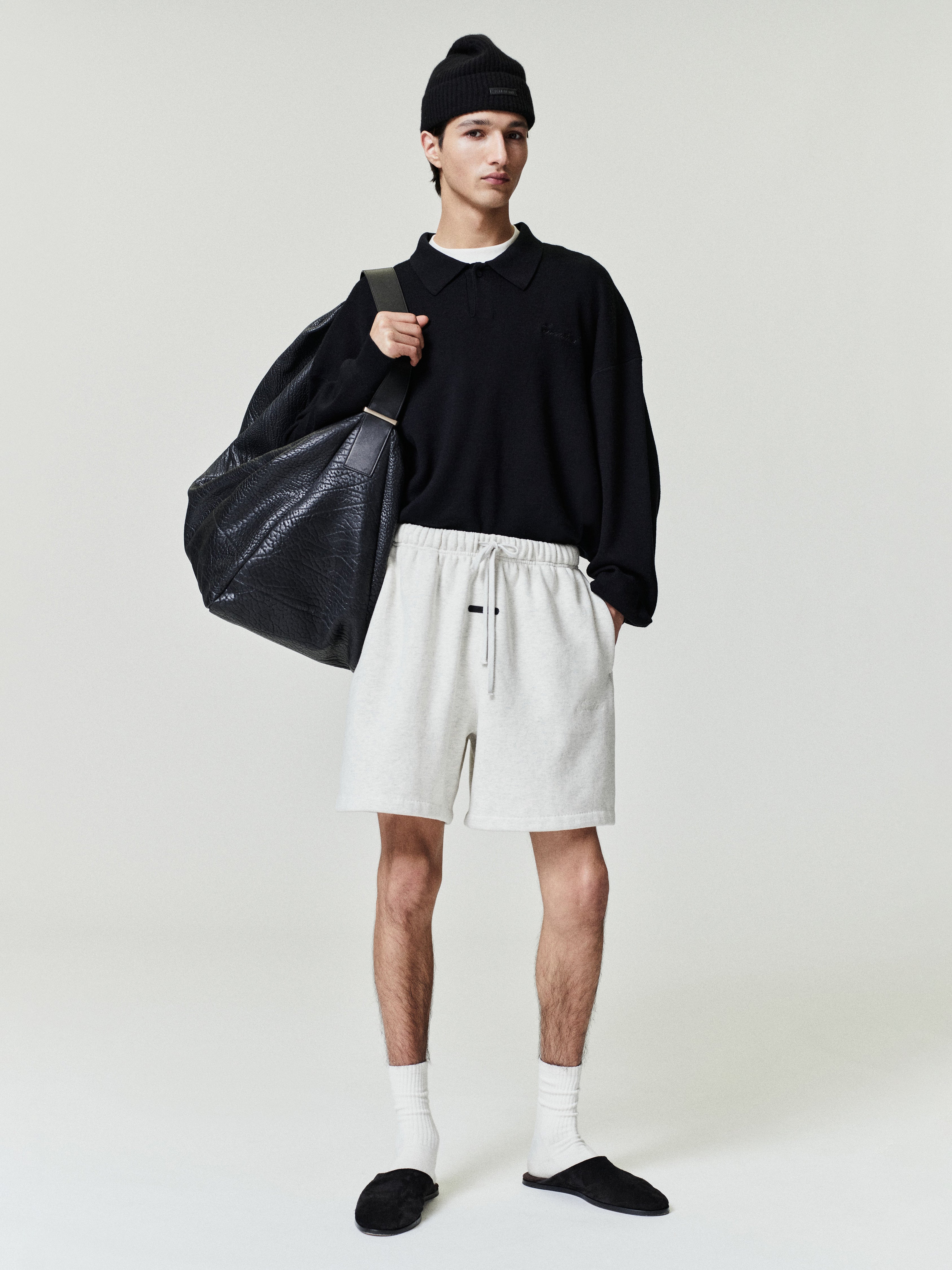 Person wearing a black sweater, white shorts, and black beanie holding a black bag on a plain background