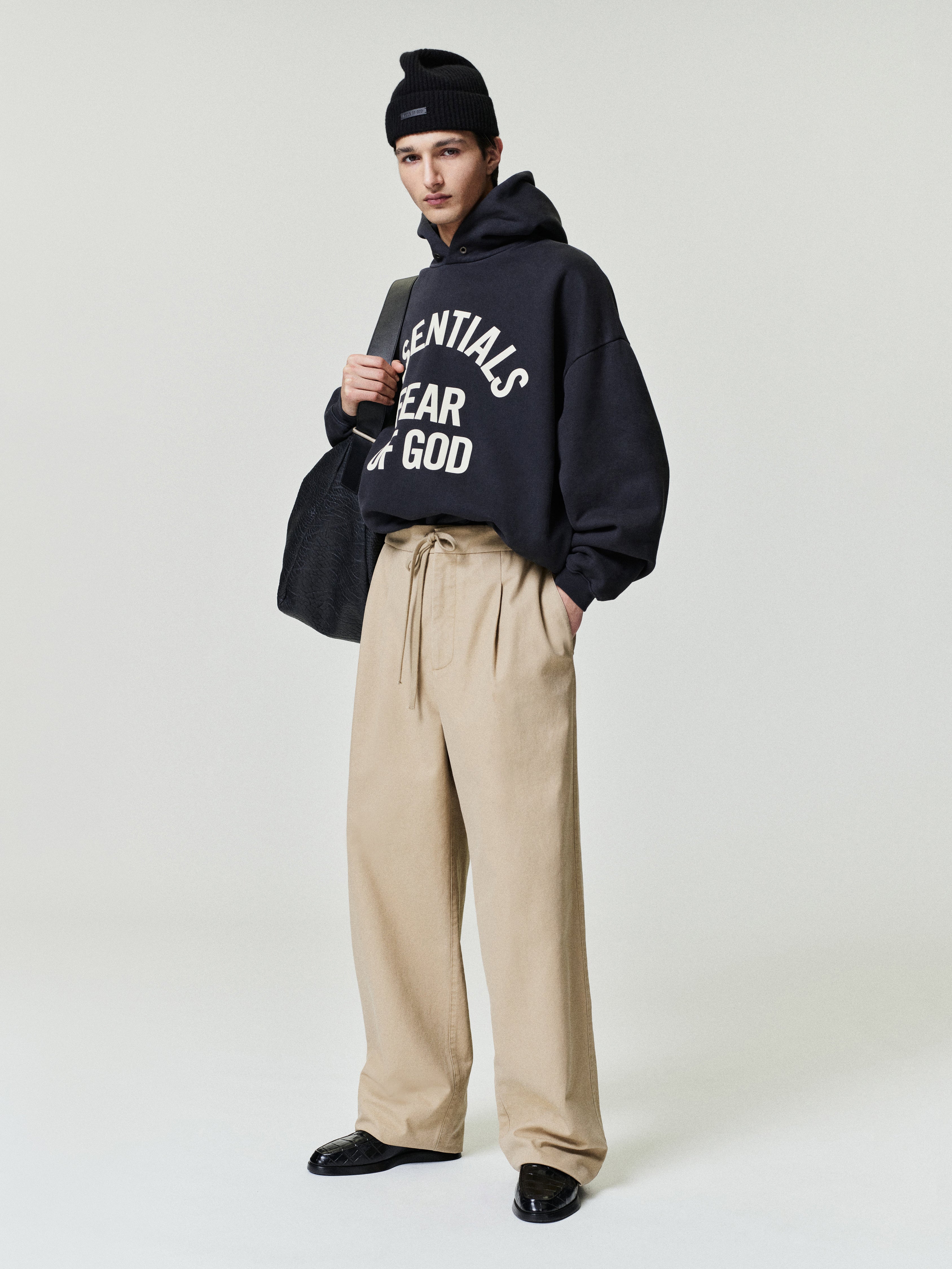 man wearing beige relaxed fit canvas pleat front pants with drawstring waistband and oversized black graphic hoodie