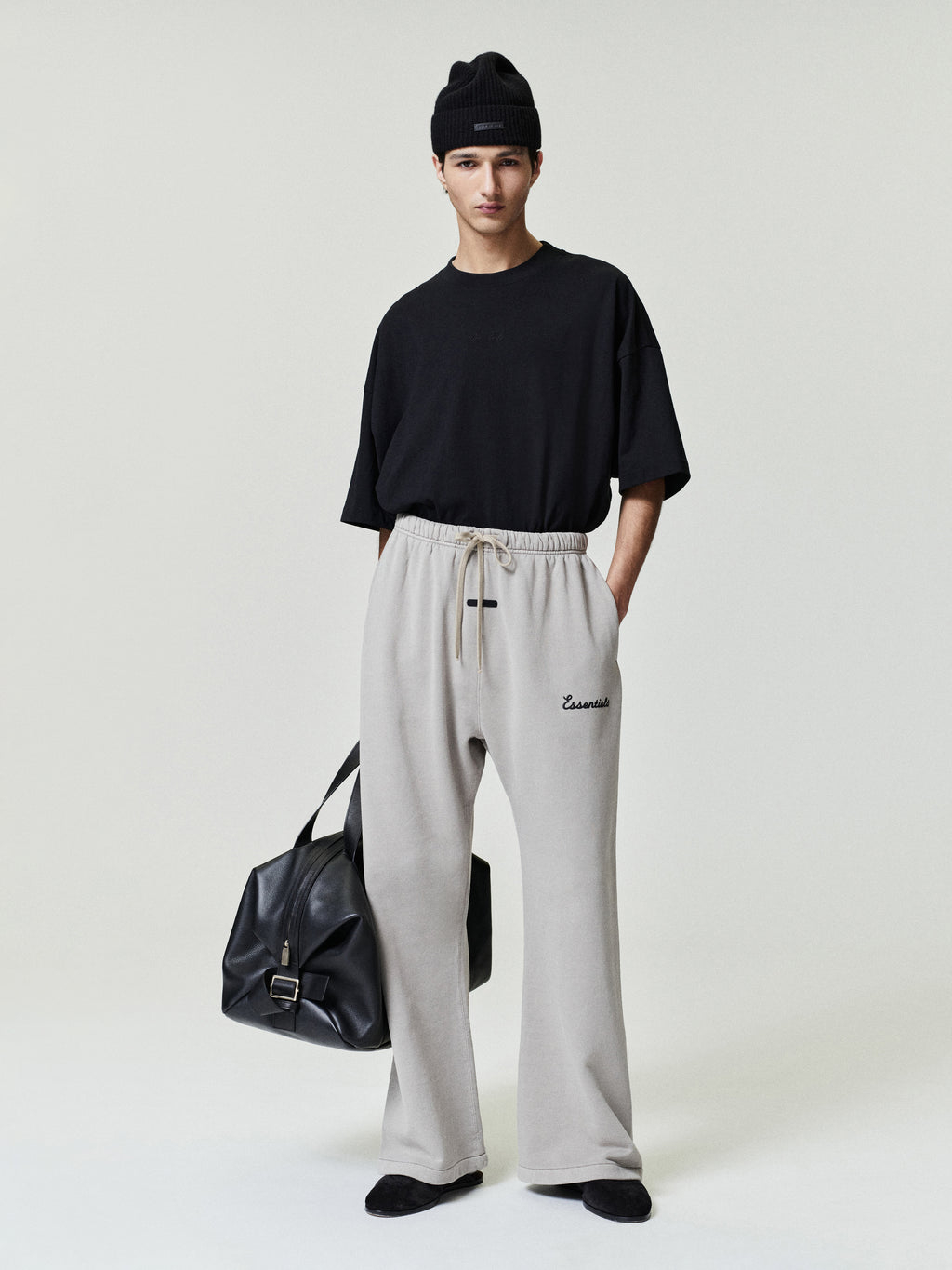 Person wearing a black t-shirt, gray sweatpants, and black beanie with a black bag on a white background
