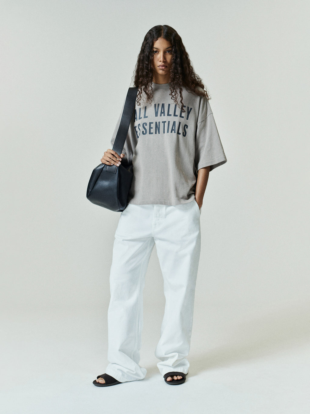 Woman wearing a Womens Merch Tee Boxy Fit in a soft taupe shade with bold navy lettering, styled with loose white pants, black slide sandals, and a black shoulder bag.