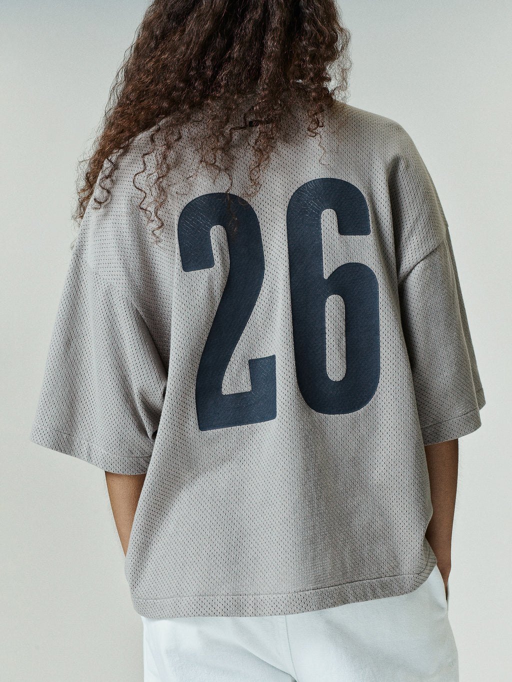 Back view of Womens Merch Tee Boxy Fit in a light neutral tone featuring a loose, breathable mesh fabric with an oversized fit and large bold black number 26 printed on the back.