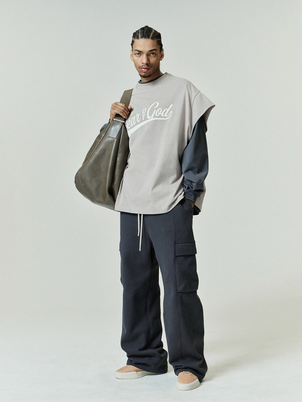 Man dressed in a layered look featuring a taupe Cut Off 90s Tee over a long-sleeve dark shirt, paired with dark cargo pants and light beige slip-on shoes, carrying a large olive tote bag.