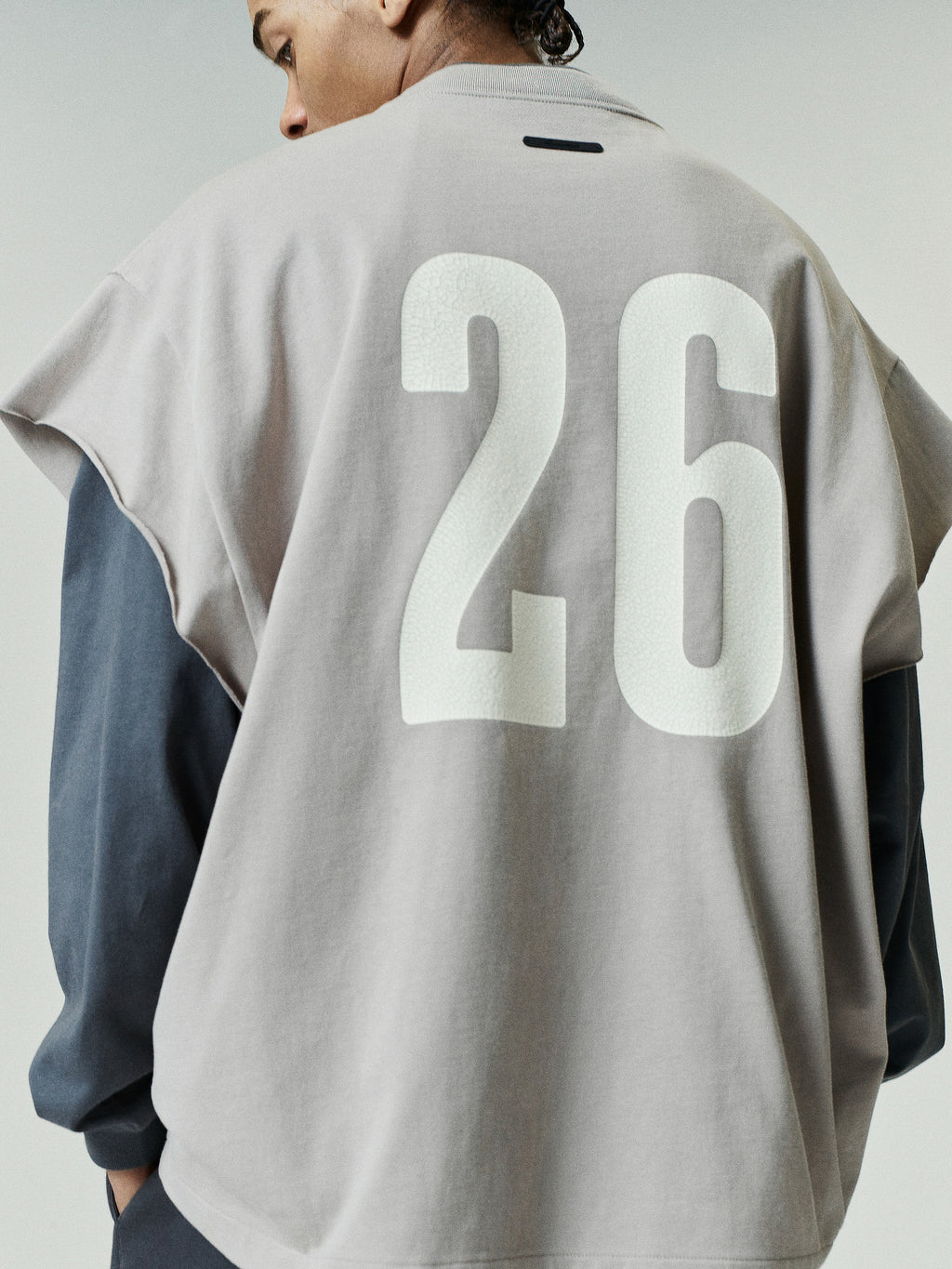 Man wearing a taupe Cut Off 90s Tee layered over a dark long-sleeve top, featuring bold white number 26 printed on the back in a relaxed casual style.