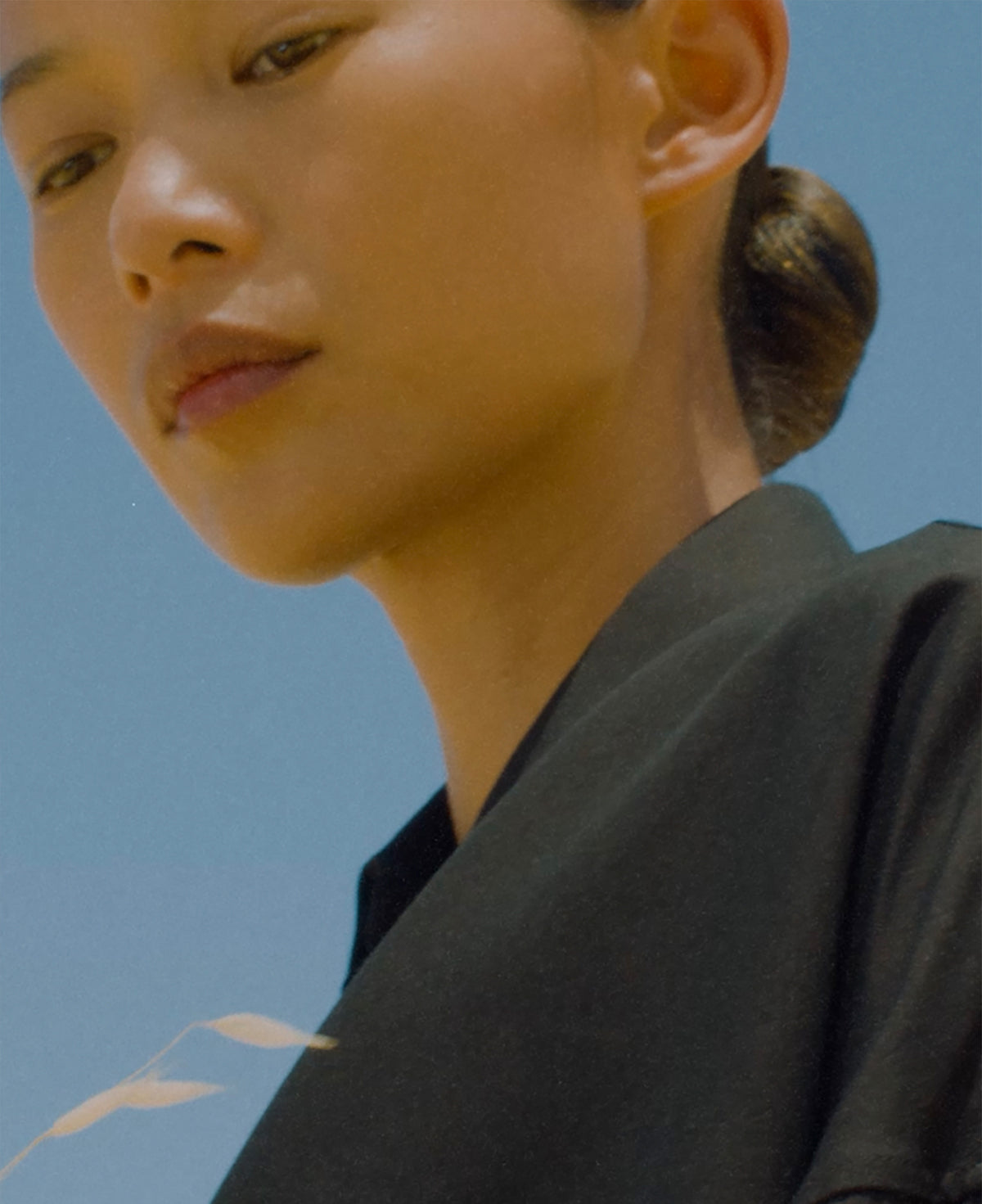 Close portrait of a woman with hair tied in a low bun wearing a black top against a clear blue sky with soft sunlight on her face