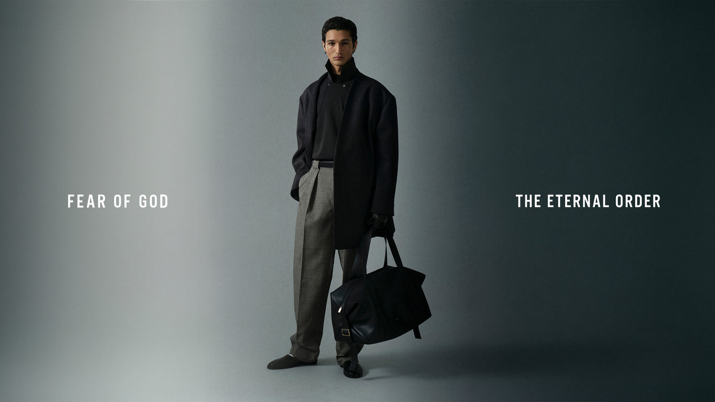 Man dressed in dark jacket and gray wide-leg trousers carrying a large black bag against gradient background